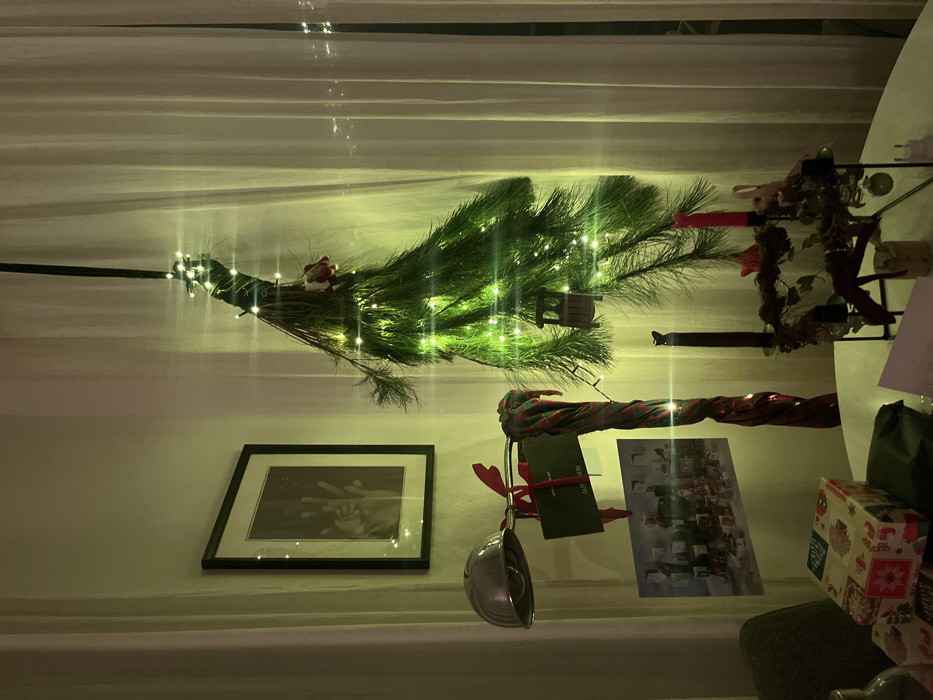 An upside-down Christmas tree with lights adorns the ceiling. Below, a framed picture hangs above a shelf displaying a Christmas card and presents. A garland of red and green fabric also rests on the shelf. The scene is lit by overhead lighting, casting a warm glow.