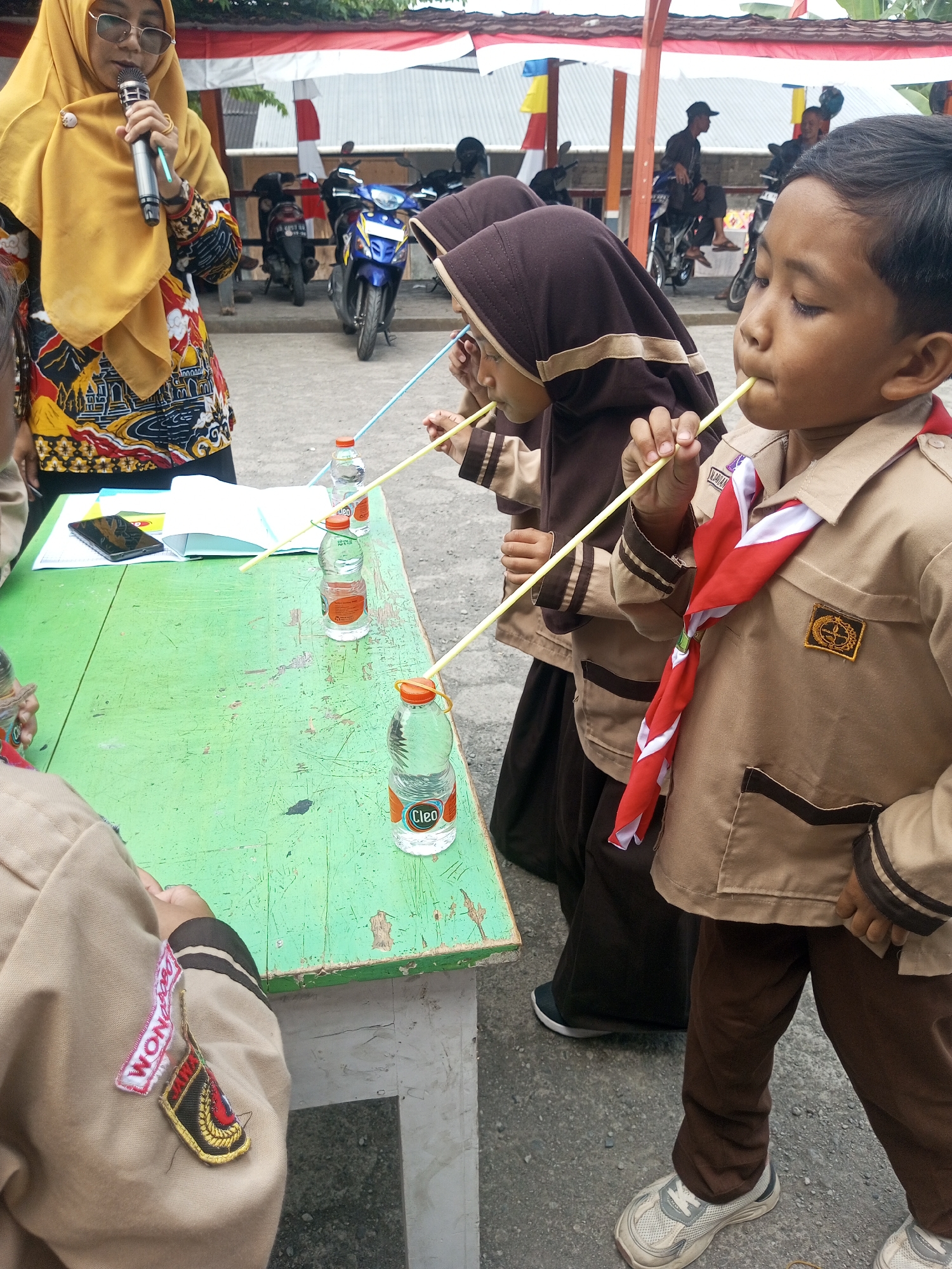 In the city of Kalibeber, Indonesia, a group of children in uniform participate in a straw-sucking game. The game involves three children, two girls and one boy, wearing tan colored uniforms with brown hijabs for the girls. The boy in the foreground is wearing a red and white scarf around his neck. They are all intently focused on sucking liquid through straws from three clear plastic bottles with orange caps arranged on a green wooden table. A woman in a yellow hijab and a patterned top stands to the left, holding a microphone, seemingly leading or supervising the activity. The table has some papers and a smartphone on it. In the background, motorcycles are parked, and a red and white flag is visible, suggesting a community event or school activity. The ground is paved with asphalt.