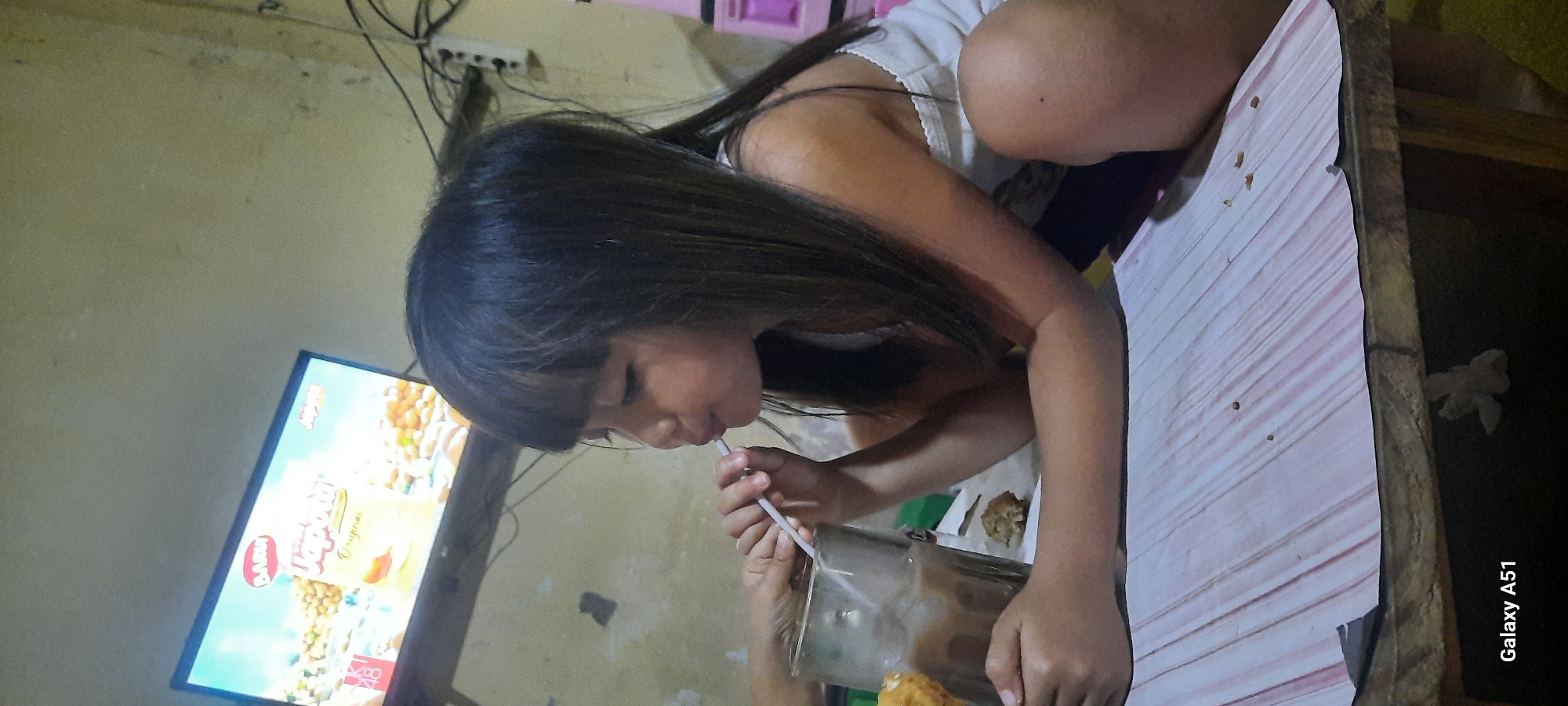 In a dimly lit room, a young girl with long dark hair is seated at a table, focused on drinking from a glass with a straw. She is wearing a white top and appears to be engrossed in her drink. To her left, a television screen displays a colorful advertisement, possibly for a food product, featuring images of what look like nuts and a cartoon character. The wall behind her is a plain, textured yellow. Electrical wires are visible running along the top of the wall, and a power strip with multiple outlets is mounted near the ceiling. The table she is sitting at has a light-colored surface with a faint pink striped pattern and appears to be made of wood. A small amount of food, possibly a snack, is on the table near her. The overall scene suggests a casual indoor setting, possibly a home. The lighting is subdued, creating a relaxed atmosphere. The camera watermark "Galaxy A51" is visible on the right side of the image.