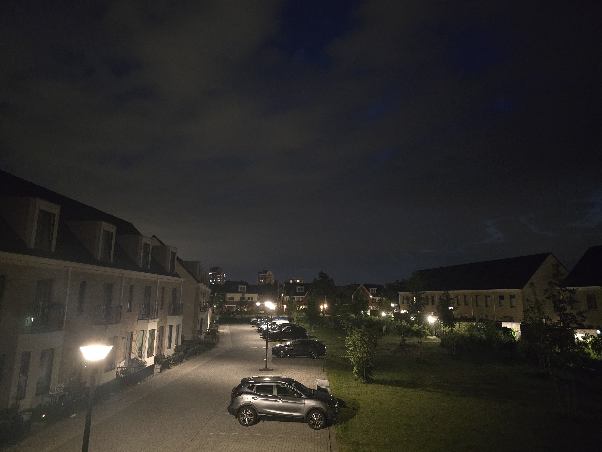 The image shows a street scene at night in Zoetermeer, Netherlands. It features a row of brick townhouses on the left side of the street, illuminated by a street lamp in the foreground. Several cars are parked along the street. On the right side, there is a grassy area with a few trees and shrubs. In the background, other buildings and street lights can be seen. The sky is dark, with scattered clouds.