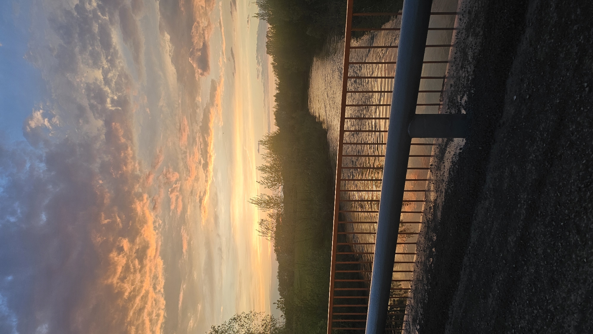 The photograph captures a sunset scene in Yangiskain, Russia, from the vantage point of a bridge or elevated structure. A river or waterway flows through a forested area, with the sun reflecting off the water's surface. The sky is filled with orange and blue clouds. A metal railing runs along the right side of the frame. The foreground shows a dark, coarse surface, possibly asphalt.