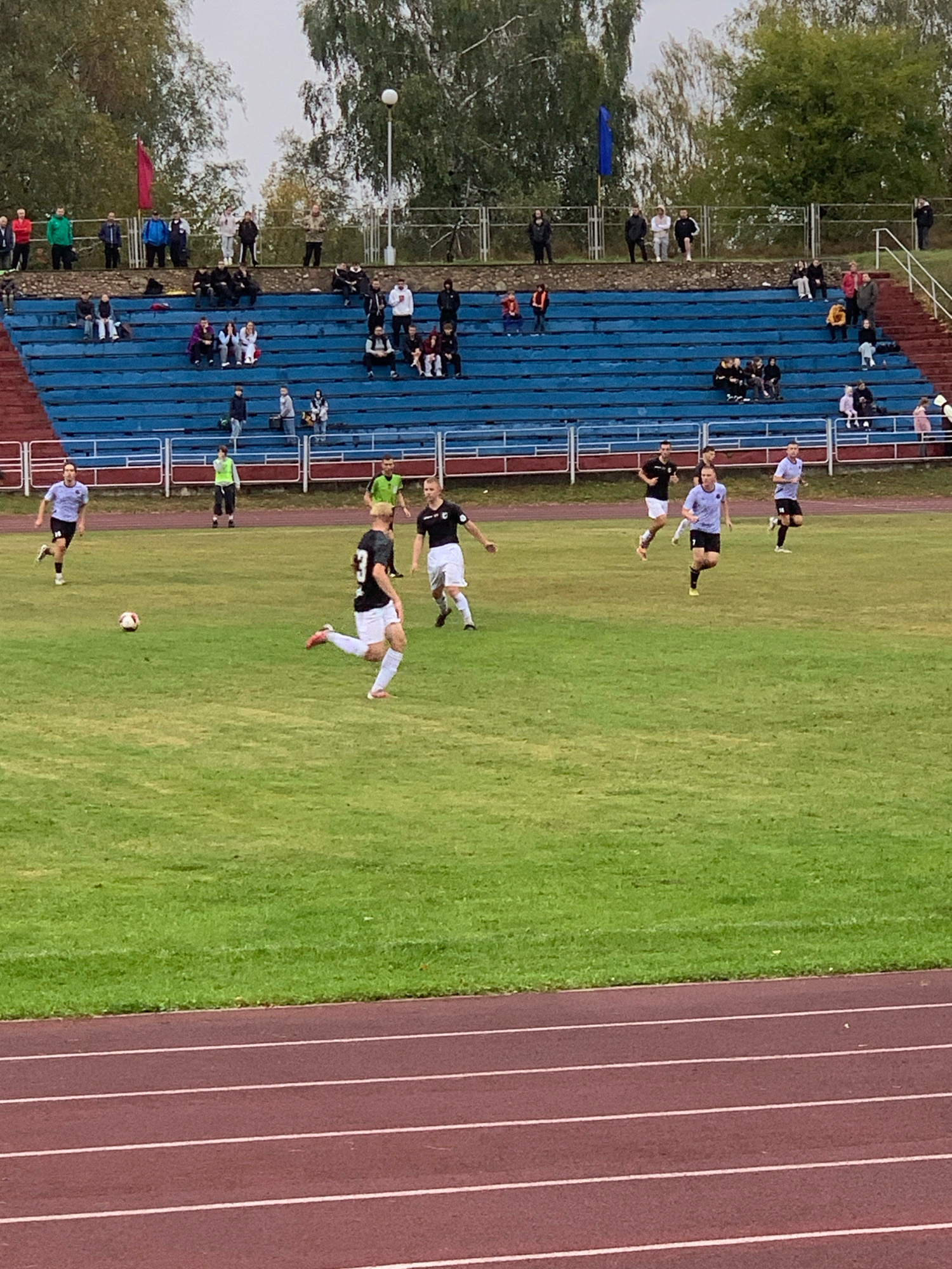 The image depicts a soccer match in progress at a stadium in Kapyĺ, Belarus. Several players are visible on the green grass field, actively engaged in the game. Some players are wearing black and white uniforms, while others are in lighter colored jerseys. A soccer ball is in motion on the field. In the background, spectators are seated in blue bleachers, observing the game. The stadium appears to be an outdoor facility with trees and a cloudy sky overhead. The scene captures a typical sporting event with players and an audience.