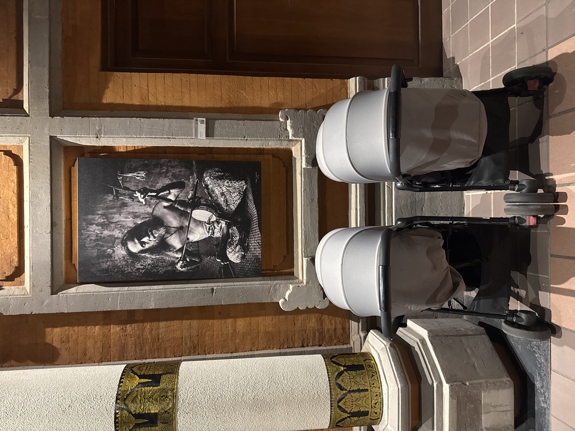 The photograph shows two empty, grey baby strollers positioned side-by-side on a tiled floor in front of a wall. Behind the strollers, a framed black and white photograph of a man playing a stringed instrument is displayed. The wall is constructed of orange bricks and has decorative elements. To the left of the strollers and the framed photograph is a large, textured white pillar with a golden decorative band near its base. To the right of the framed photograph, a dark wooden door is visible. The lighting in the scene is warm and creates shadows on the floor. The image appears to be taken indoors, possibly in a gallery or public space. The location is identified as Kuta Selatan, Indonesia.
