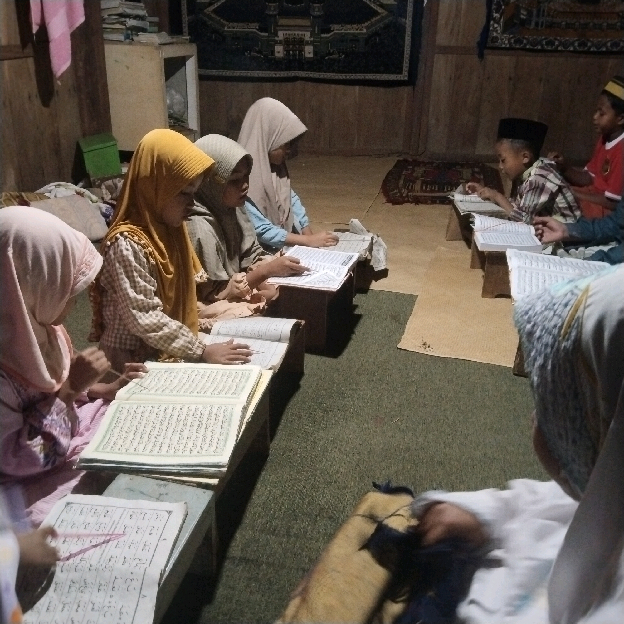 In Banyubunih, Indonesia, a group of children are gathered indoors for what appears to be a Quranic study session. The room is dimly lit, with wooden walls and a rug on the floor. Several children, some wearing hijabs, are seated on the floor around low wooden stands, each holding an open book, likely the Quran. One child in the foreground is wearing a pink hijab and reading from a book. To their right, another child in a yellow hijab is also reading. Further back, more children are engaged in similar activities. Two boys in the background are also reading from books. The atmosphere suggests a focused and educational environment.