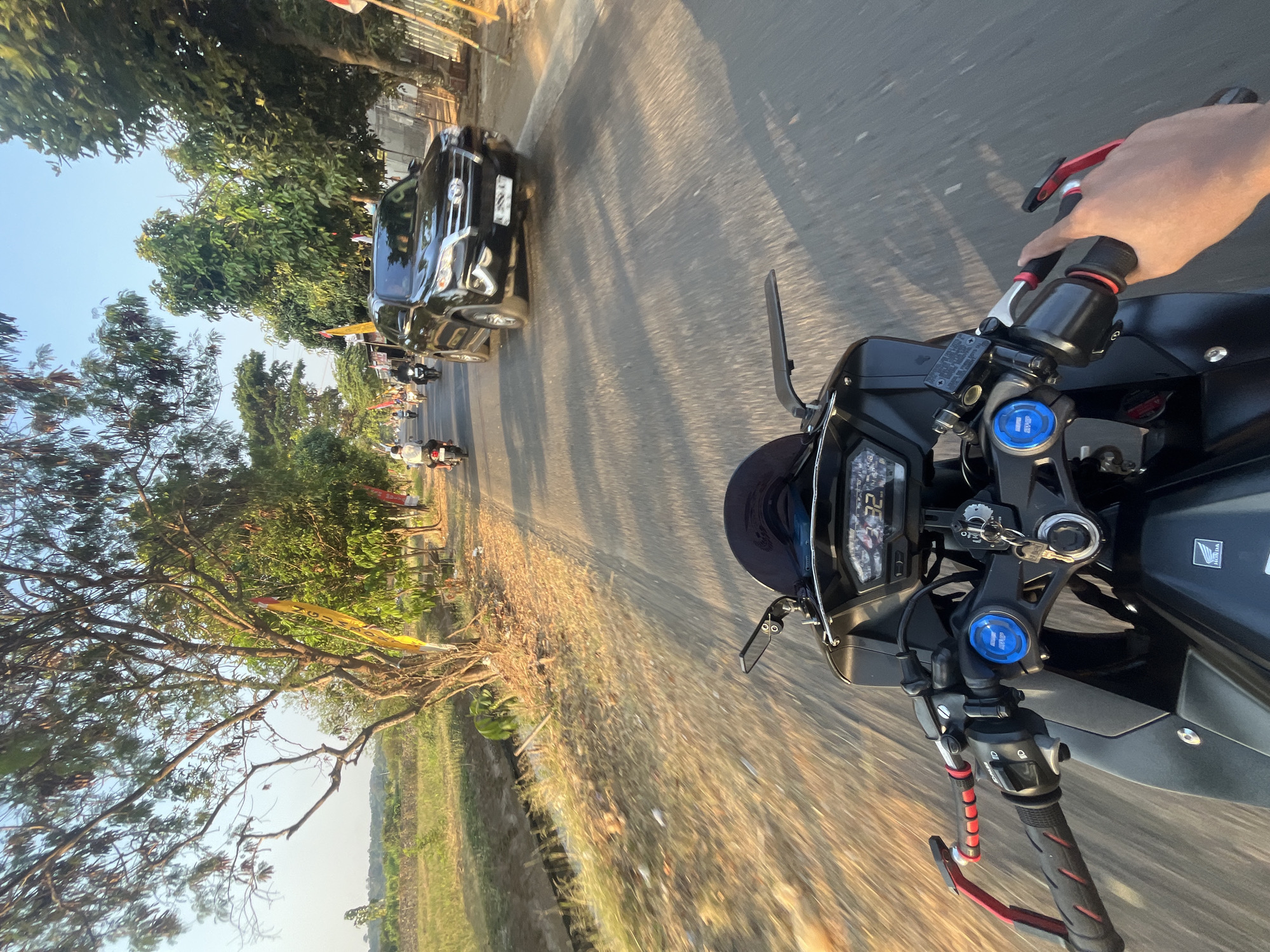 The image depicts a first-person perspective of someone riding a black motorcycle on a paved road. The motorcycle's handlebars, instrument cluster, and part of the front fairing are visible in the foreground. The road appears to be in a rural or semi-rural area, with vegetation and a ditch along the side. Several other motorcycles and a black SUV are also present on the road, suggesting traffic. Flags, possibly for a local event or celebration, are visible along the roadside. The time of day appears to be late afternoon or early evening, indicated by the warm, golden sunlight and long shadows. The location is identified as Pecangaan, Indonesia.