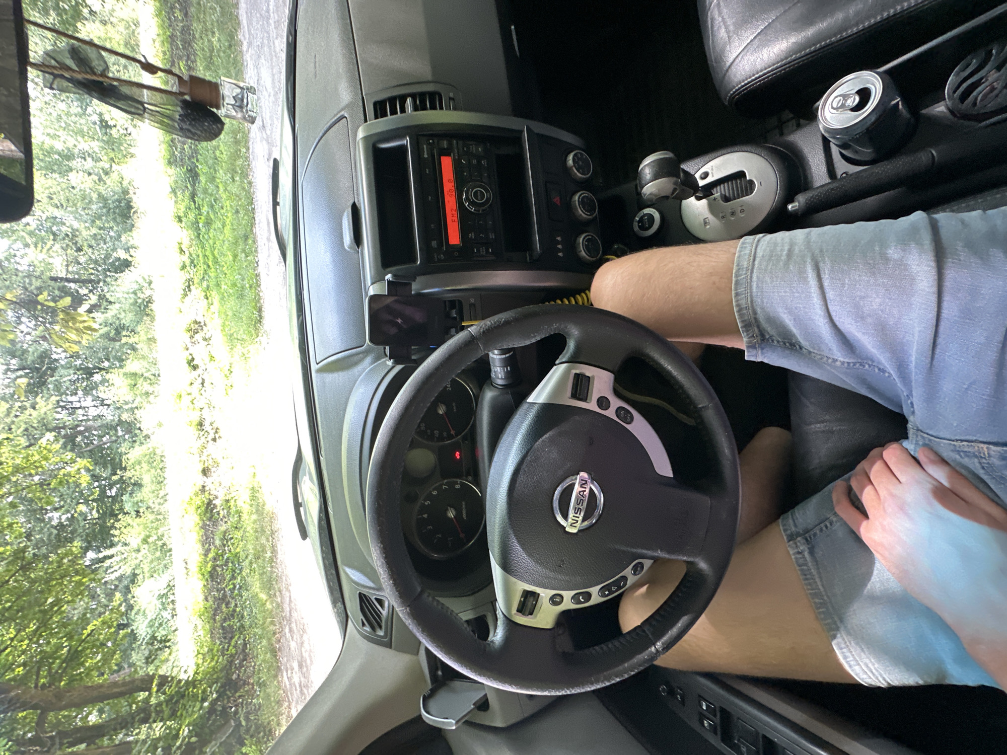 The image shows the interior of a Nissan car, possibly a Qashqai, parked on a dirt road surrounded by trees and greenery, indicative of a rural setting. The dashboard features a steering wheel with the Nissan logo, radio controls, and a gear shifter. A smartphone is mounted on the dashboard, and a can of soda is visible in the center console. The driver's legs are visible, wearing denim shorts. The scene appears to be taken during the daytime, with sunlight filtering through the trees. The location is identified as Shatava, Ukraine, suggesting a recreational or travel context. The overall impression is that of a paused moment during a road trip in a natural environment.