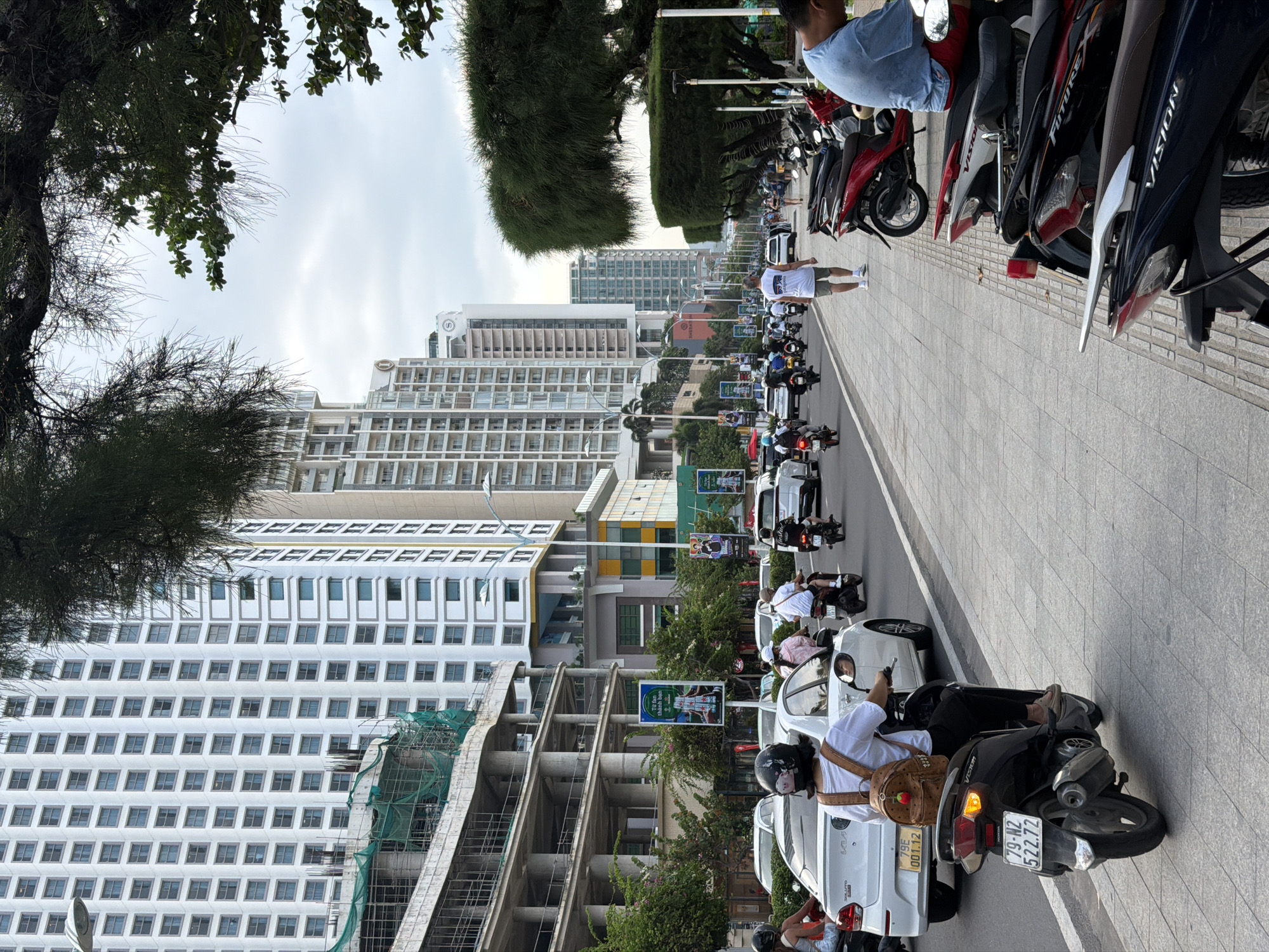 This daylight image captures a bustling street scene in a Vietnamese city, likely Nha Trang, characterized by a mix of modern high-rise architecture, dense traffic, and street-level activity.
In the foreground, a wide, tiled sidewalk is lined with numerous parked motorcycles, predominantly Honda models like "FUTURE" and "VISION." Several people are present; one individual in a light blue shirt is seated on a motorcycle on the far right, partially obscured. Another person in a white t-shirt and shorts walks on the sidewalk further back.
The street itself is busy with traffic, dominated by motorcycles and scooters, with a few cars interspersed. Riders are seen wearing helmets, navigating the road. Green foliage from trees lines both sides of the street, with lush, dense trees framing the upper left and extending along the thoroughfare.
In the midground and background, an urban landscape of tall, contemporary buildings rises, many appearing to be hotels or residential high-rises. A prominent "Sheraton" logo is visible on top of one of the buildings. Near one of the structures, scaffolding and green construction netting indicate ongoing development. Billboards with green backgrounds and white text, possibly advertisements for "thành hơn" (making it better/more successful) and "Vinpearl," are positioned along the street.
The sky overhead is overcast, suggesting a cloudy day but with ample natural light. Visible text on license plates includes "79-N2 522.72" on a black scooter and "79G1 001.72" on a white car, both common formats in Vietnam. One motorcycle rider in the foreground carries a small, strawberry-patterned backpack. The overall scene depicts a vibrant and active urban environment.