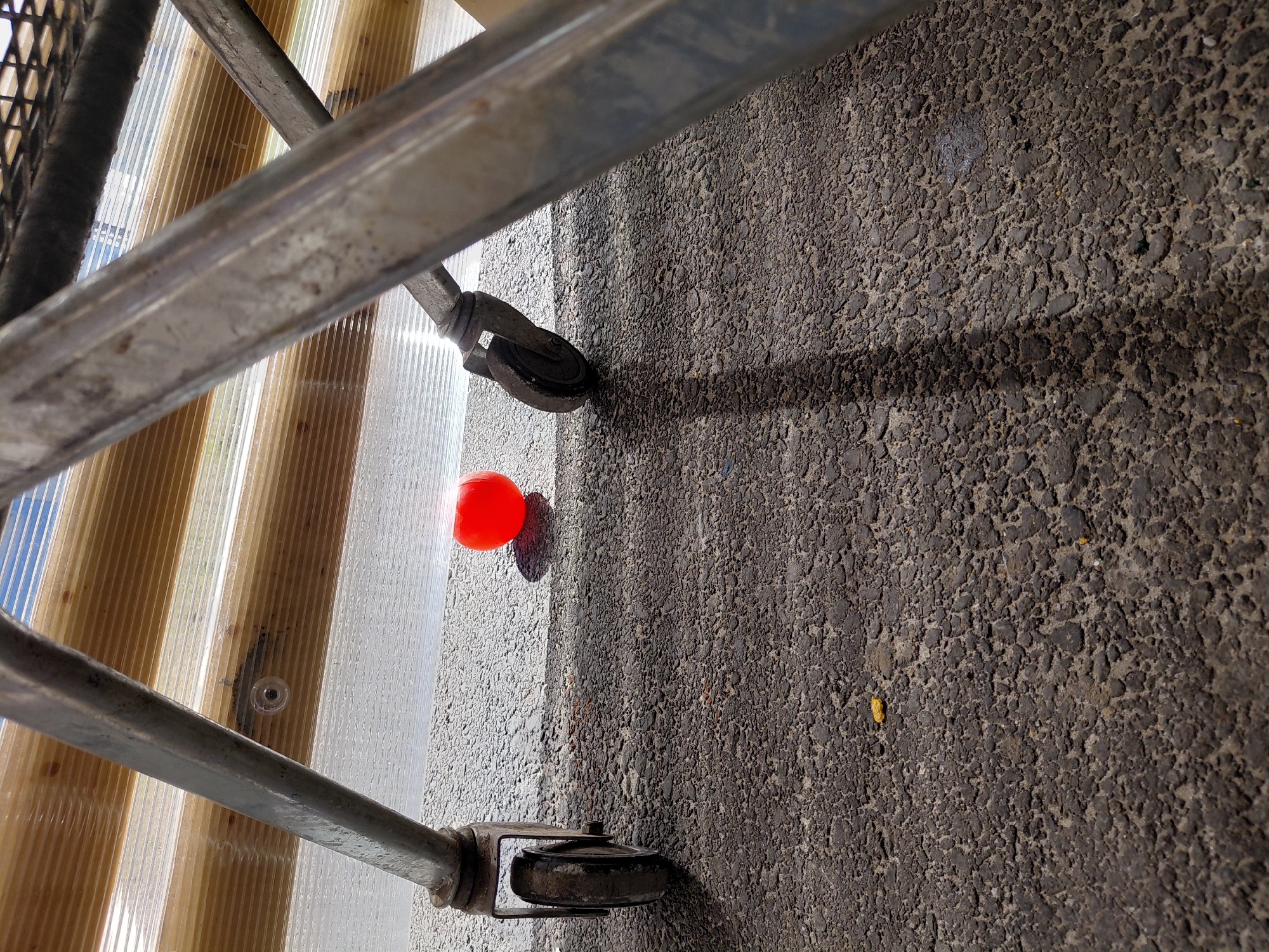 The image displays an outdoor, slightly overhead view of a ground-level scene, illuminated by strong, direct sunlight. A metallic structure, possibly part of a cart, trolley, or light scaffolding, crosses the frame diagonally from the top-left to the bottom-right. This structure appears to be made of two parallel, worn, and somewhat discolored metal bars. Attached to the lower ends of these bars are two dark, scuffed casters or wheels, one visible in the upper-right corner and another in the lower-left, indicating the object's mobility.
To the right of the metallic structure, a translucent, corrugated panel extends vertically. This panel, likely plastic or polycarbonate, features alternating sections of clear/white and a light amber or brown tint. A small, circular fastener is visible embedded within one of the clear corrugated sections.
The left side and the majority of the background are covered by a dark, coarse, and highly textured ground surface, characteristic of asphalt or rough concrete. A prominent, sharp shadow cast by the metallic structure stretches across this ground, creating a distinct boundary between a brightly lit, lighter-toned strip of ground and a larger, darker shaded area. A tiny, bright yellow speck is noticeable within the shaded ground.
Near the upper-center of the image, a vibrant red, spherical object, resembling a small ball, rests at the intersection of the corrugated panel and the ground, casting a short, distinct shadow. The bright lighting and sharp shadows strongly suggest a clear, sunny day, likely mid-day. No people or visible text are present in the scene, which is located in Nadym, Russia.