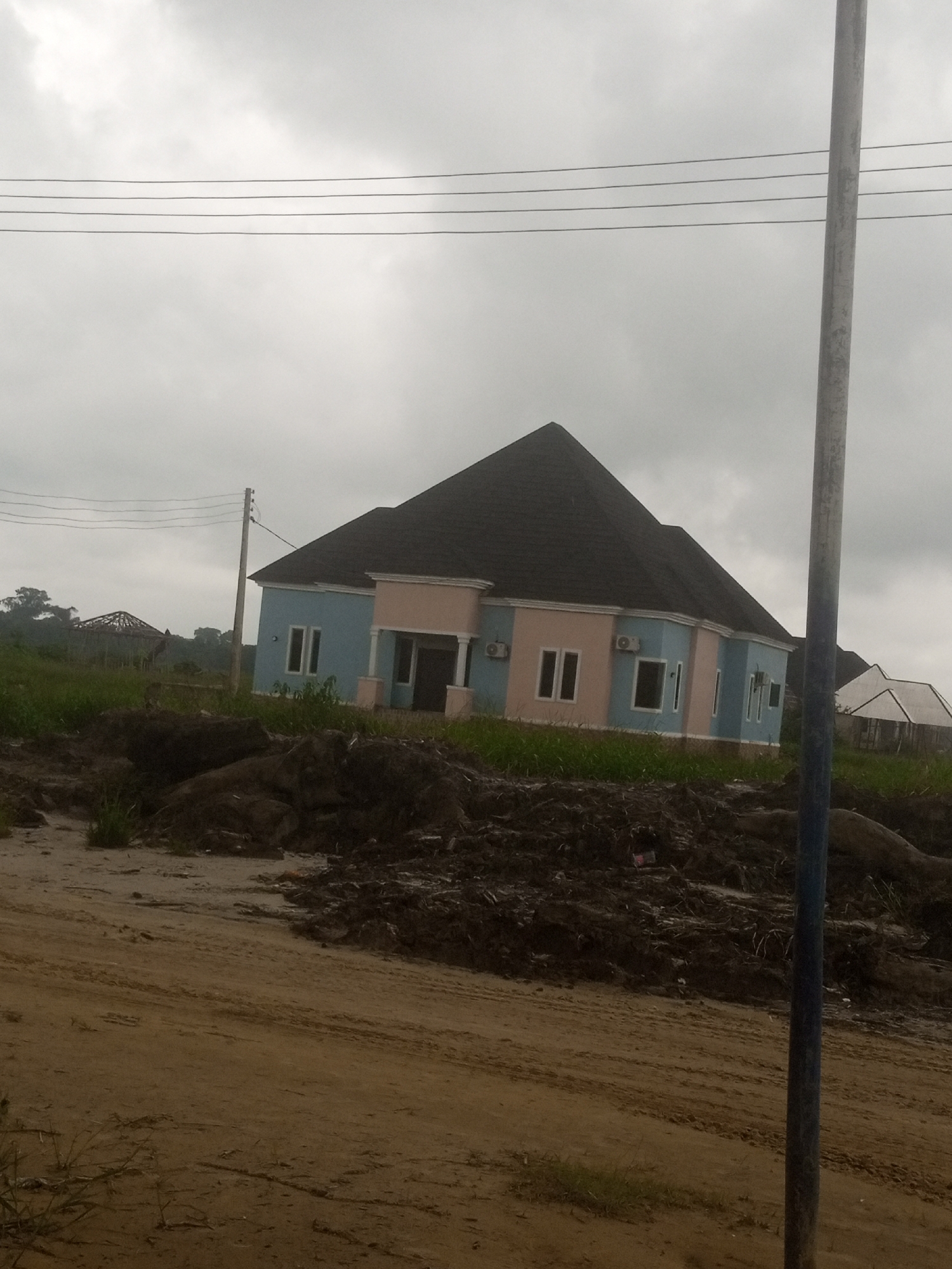 The image shows a blue and pink house with a dark roof, situated in an undeveloped area. The house has several windows and what appear to be air conditioning units mounted on the walls. In the foreground, there is disturbed earth and a dirt path. In the background, there are more buildings under construction, including a circular structure with a roof frame, and some vegetation. Overhead, power lines stretch across the cloudy sky. A grey pole with blue markings stands prominently in the foreground on the right side of the image. The scene is consistent with a construction site in a rural or semi-developed area, likely in or near Benikrukru, Nigeria. There are no people visible in the image.