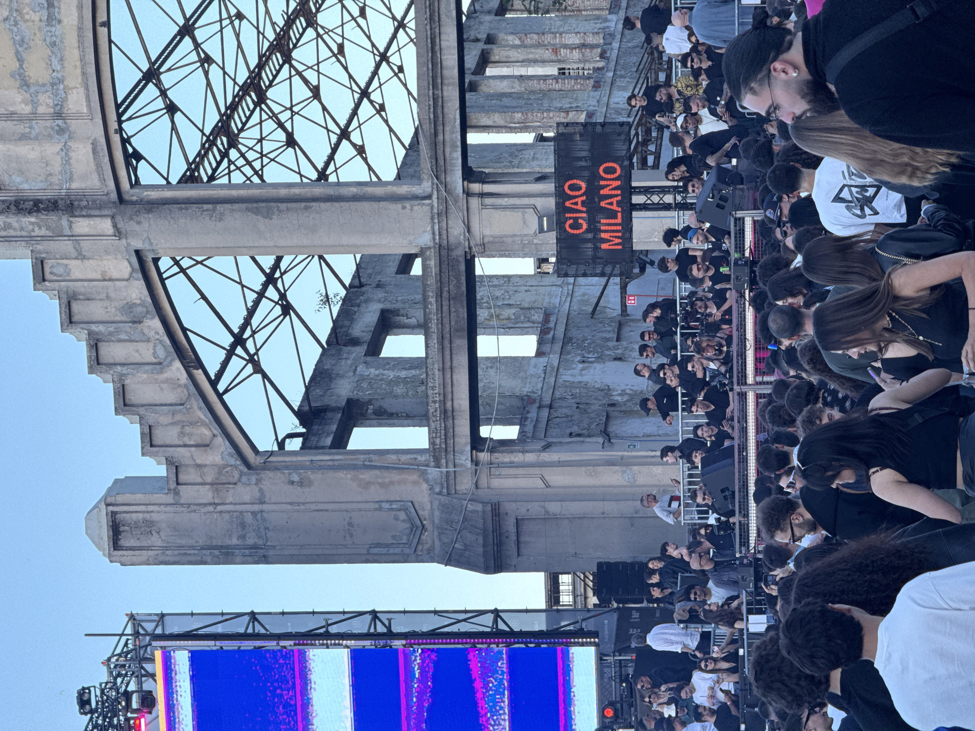 The image depicts a crowded outdoor event in Milan, Italy, likely a fashion show or concert, indicated by the "Ciao Milano" sign displayed in red letters. An audience of mostly young people, many dressed in black, are looking towards a stage. The setting appears to be within or near a historical or industrial ruin, with a skeletal concrete structure and exposed metalwork visible above the crowd. A large screen displaying bright blue and magenta patterns is positioned below the ruined structure.