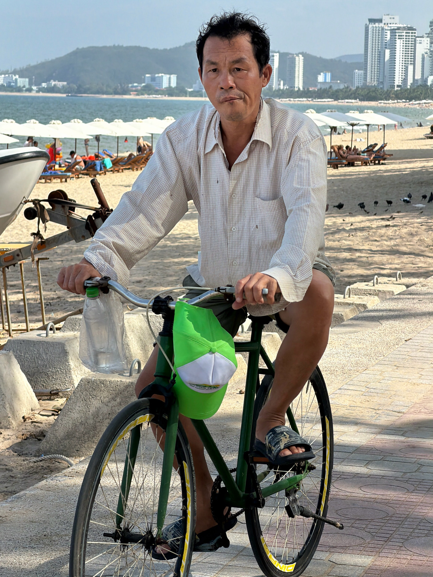 A middle-aged East Asian man wearing a plaid button-down shirt and shorts is sitting on a bicycle on a paved walkway next to a sandy beach. The man is looking towards the camera with a neutral expression. The bicycle has a green frame and a green baseball cap with a white brim hanging from the handlebars. A plastic bottle of water is attached to the handlebars. In the background, there is a beach with lounge chairs and umbrellas, the ocean, and a city skyline with tall buildings. The scene appears to be in Nha Trang, Vietnam, given the presence of the beach, ocean, and city architecture characteristic of the area.