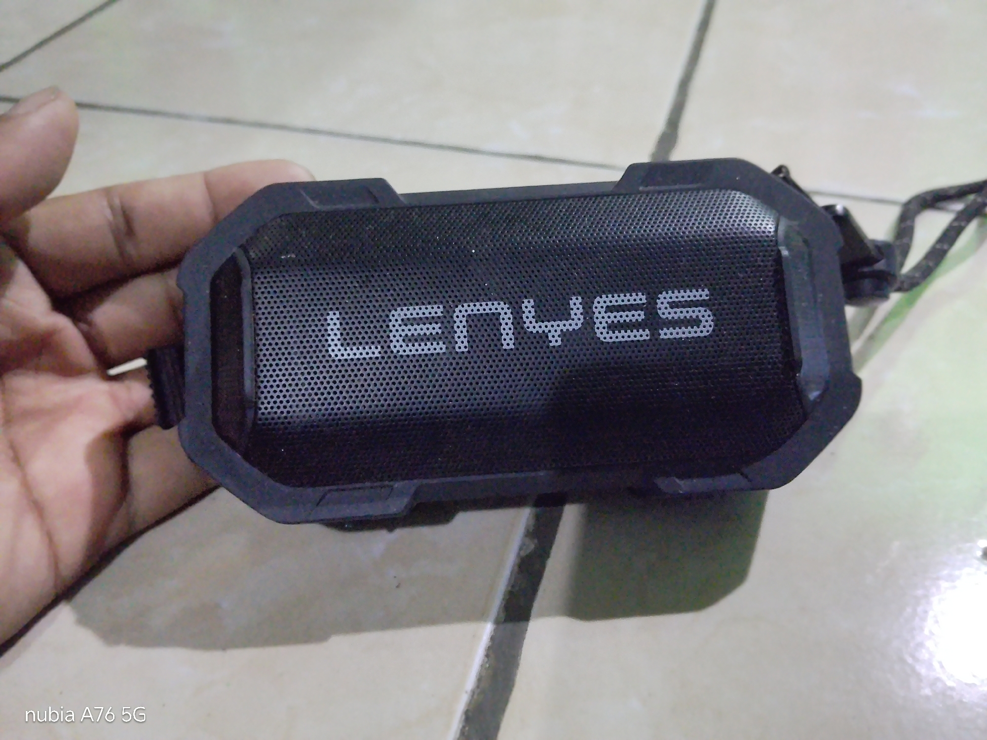 A close-up shot shows a black portable speaker with the brand name "LENYES" embossed on its mesh front. A person's hand is visible, holding the speaker. The speaker is rectangular with rounded edges and appears to be made of a durable material, possibly rubberized. A black lanyard is attached to one of the speaker's sides. The speaker rests on a tiled floor. The timestamp "nubia A76 5G" is visible in the lower-left corner of the image, indicating the device used for capture. The scene appears to be indoors, with the tiled floor suggesting a domestic or commercial setting.