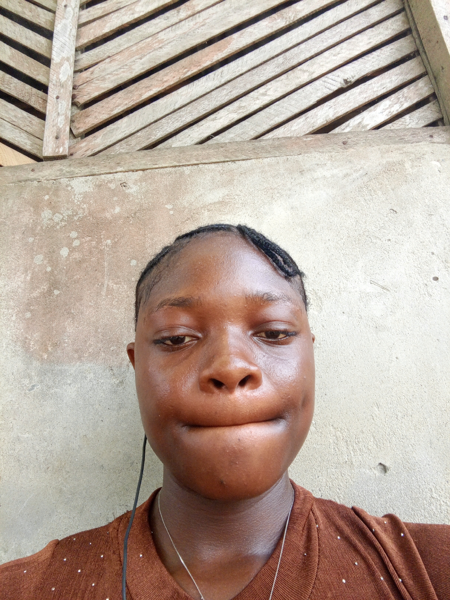A close-up selfie of a young woman is centered in the frame. She has dark brown skin, short black braids, and is wearing a brown shirt with small, white dots. A thin, silver necklace is visible around her neck, along with a black earbud wire hanging down. Her lips are slightly pursed, and her expression is somber. The background consists of a weathered wall, possibly concrete or plaster, with visible staining and discoloration. Above the wall, there is a section of wooden planks arranged diagonally, suggesting a partially constructed or dilapidated structure. The setting appears to be outdoors in Mbribit, Nigeria, considering the query context.