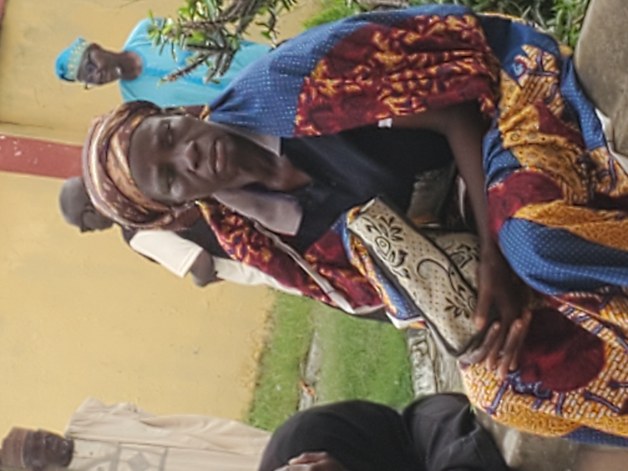 The image portrays an outdoor scene in Mararaba, Nigeria, featuring multiple individuals against a plain yellow wall. In the foreground, a dark-skinned adult, likely female, is seated on a concrete structure. She wears a dark blue polo-style shirt, a brown and gold patterned headwrap, and is draped in a wrapper or shawl featuring distinct patterns in blue, red, and yellow/gold. Her hands are clasped over a beige or cream-colored handbag with a dark, intricate pattern, resting in her lap. Her facial expression is neutral, and she looks slightly to the right of the frame. In the mid-ground, standing against the yellow wall, are several other individuals. From the left, an older adult male with a white beard is visible, dressed in a light-colored traditional Nigerian garment and cap. Next, a partially obscured figure with dark skin wears sunglasses. To the right of the seated individual, an adult male wears a white t-shirt and a dark vest. Further to the right, an older adult male wears a light blue traditional outfit, a patterned blue cap, and glasses, partially obscured by green foliage. The ground is a mix of green grass and concrete.
