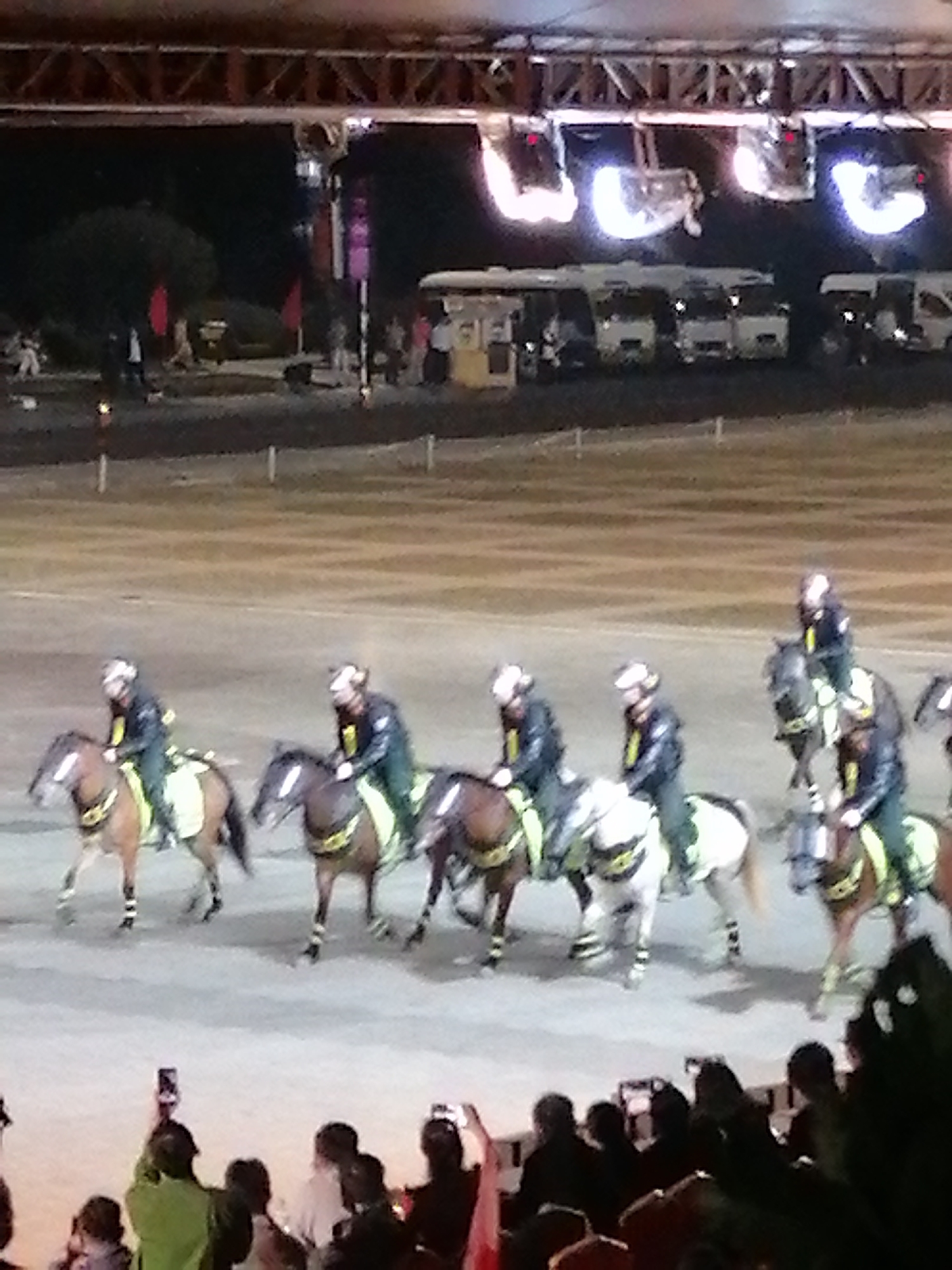 This media file depicts an outdoor equestrian event at night, likely a performance or parade. A group of riders on horseback are moving across a sandy or dirt arena. The riders are dressed in matching dark uniforms with lighter-colored trim and helmets. Their horses are adorned with matching saddle blankets and some appear to have decorative elements on their legs. The riders are positioned in a line, suggesting a coordinated movement or display.
In the background, large vehicles, possibly buses or tour coaches, are parked. Above the vehicles, a structure with lights is visible, suggesting a stage or covered area, illuminating the scene. Some of the lights are very bright, creating a hazy effect. Red flags are planted in the ground in the distance, adding a splash of color to the night scene.
In the foreground, a crowd of people is gathered, many holding up cell phones to capture the event. The presence of the crowd and the staged performance indicate a public gathering. The time of day is clearly night, as evidenced by the artificial lighting and the dark sky. The overall atmosphere appears to be one of excitement and entertainment. The location cues, such as the type of vehicles and the general setting, are consistent with Da Lat, Vietnam, known for its entertainment and event venues. There is no visible text in the image.