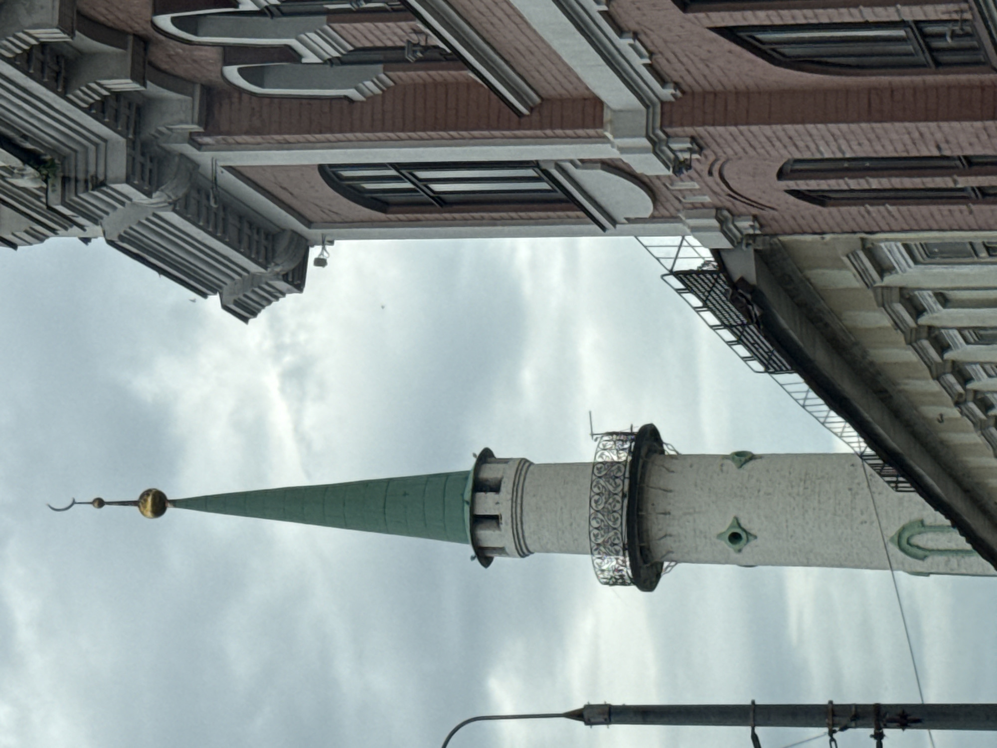 This is a photograph of a minaret with a green spire, topped with a golden orb and a crescent moon, standing against a cloudy sky. To the right of the minaret is a brick building with decorative white trim and arched windows. The minaret is tall and cylindrical, made of light-colored stone, with a decorative metal balcony partway up. Below the balcony, there are two small, diamond-shaped green windows. The brick building appears to be older, with a more ornate architectural style. The overall impression is of a city scene, with the minaret likely part of a mosque or religious complex. This scene is located in Kazan, Russia, which has a rich history of Islamic architecture.