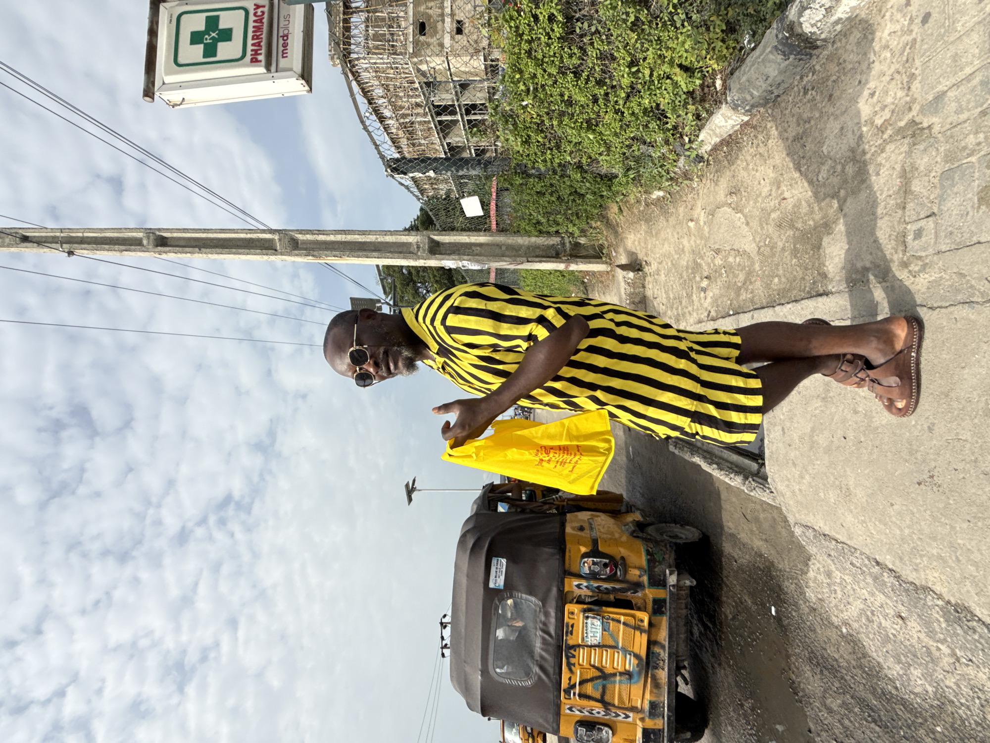 A medium outdoor shot shows a dark-skinned man, wearing a yellow and black striped short-sleeved shirt, brown sandals, and sunglasses. He is standing on a concrete sidewalk next to a yellow auto rickshaw taxi. He is holding a yellow bag in his right hand.
The background reveals a pharmacy sign, an unfinished building, and lush greenery. The sky is partly cloudy. The location is in Ado, Nigeria.
