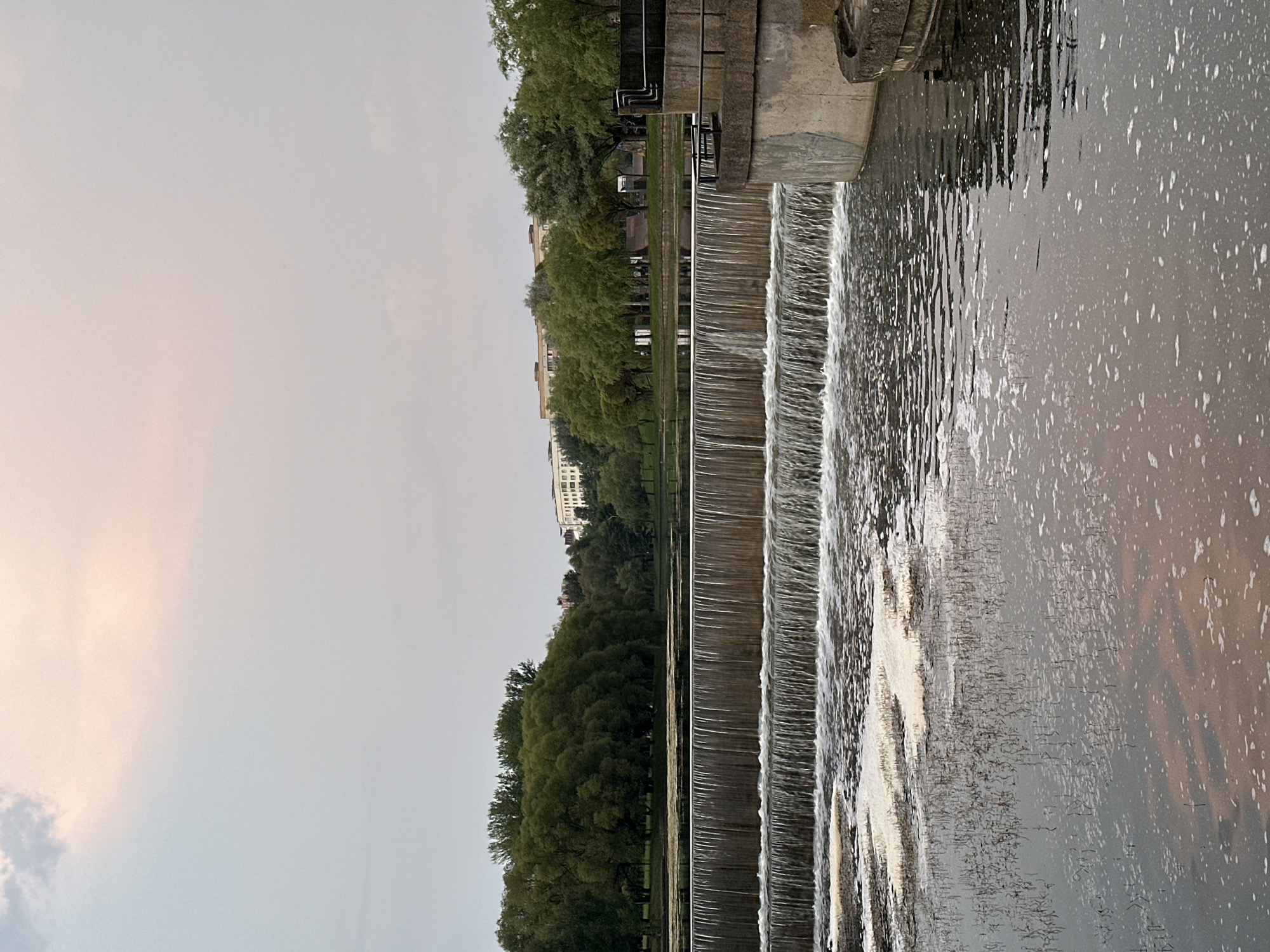 An outdoor, eye-level, medium shot captures a tiered waterfall flowing into a body of water, set against a backdrop of trees and buildings. The sky above is overcast with subtle hues of pink and pale blue, suggesting either sunrise or sunset. The waterfall, constructed from concrete or stone, cascades in multiple levels, creating a foamy white texture at its base. The water in the foreground is murky and reflects the muted colors of the sky and surrounding foliage. On the right side of the frame, a concrete structure with a railing, part of the waterfall's support system, is visible. In the distance, a line of lush green trees forms a natural barrier, behind which several light-colored buildings rise. The overall scene depicts a tranquil urban park or waterfront area. There are no people or animals present in the image.