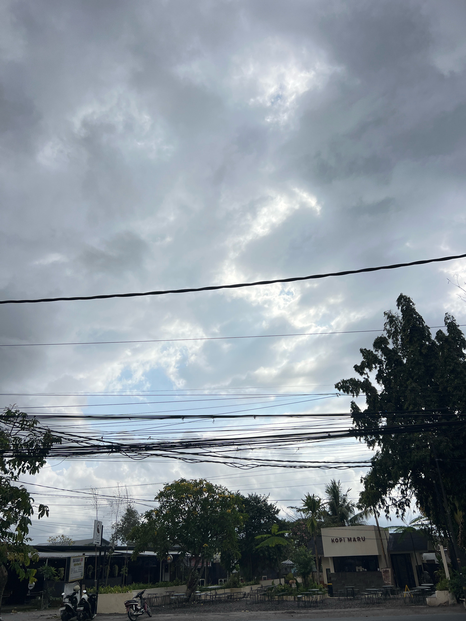 The image captures a cloudy sky over a street scene in Denpasar, Indonesia, with a building labeled "KOPI MARU" and several motorbikes parked. Overhead, a complex network of power lines crisscrosses the frame, casting shadows on the ground and obscuring parts of the sky. Lush green trees, including a tall, dense one on the right, flank the street and the building. Several tables and chairs are set up in front of the establishment, suggesting it's a cafe or restaurant. The overall atmosphere is muted due to the overcast sky, with natural light dominating the scene.