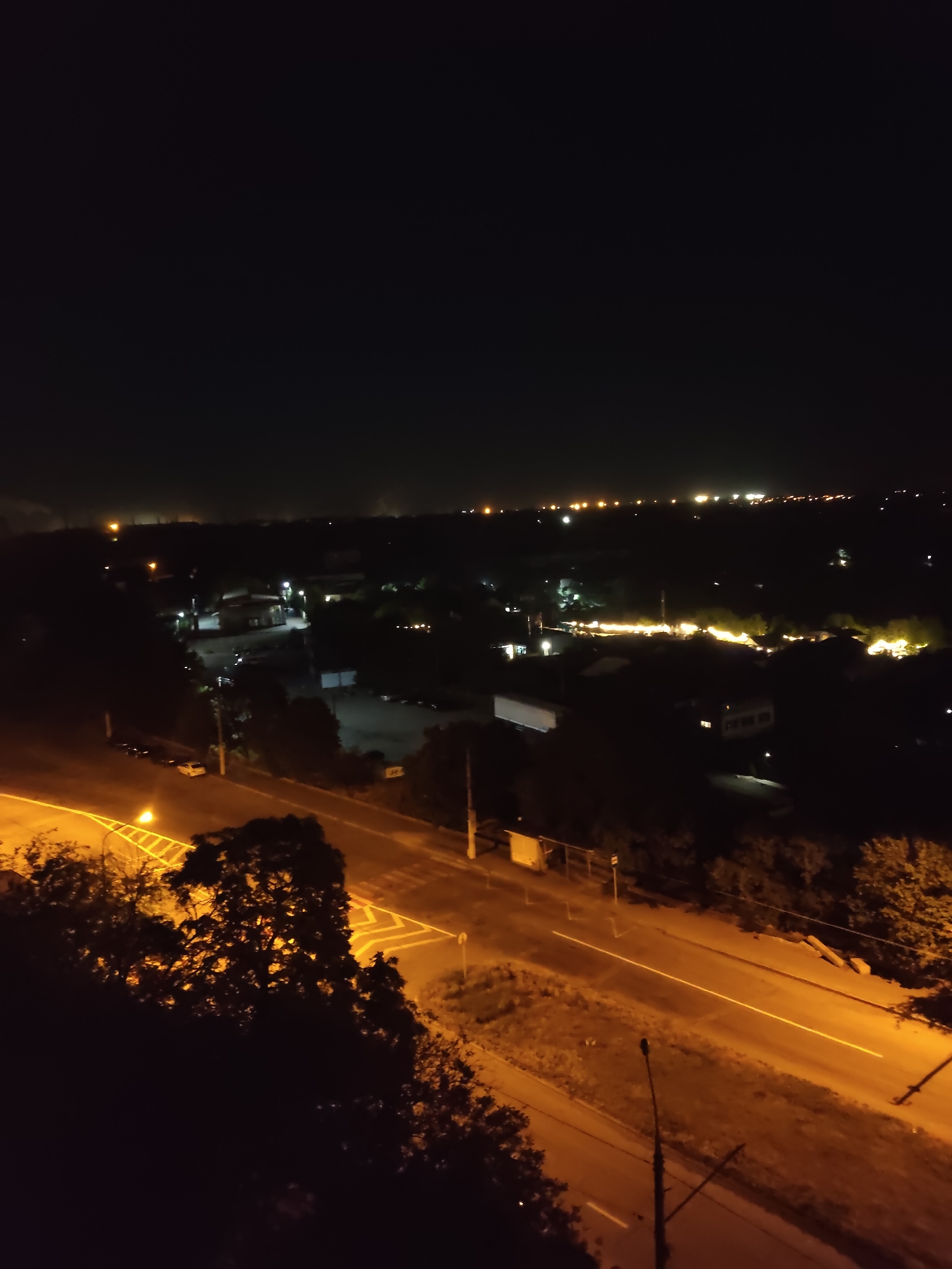 This is a nighttime aerial view of a semi-urban or industrial area in Zaporizhzhia, Ukraine. The foreground is dominated by dark trees and foliage, with a portion of a sloping, tree-covered hill. A road with yellow and white markings runs through the middle ground, illuminated by streetlights. Several vehicles are visible parked along the side of the road in the distance. The background shows a spread of buildings and lights, suggesting a town or industrial zone. The sky is dark and featureless.