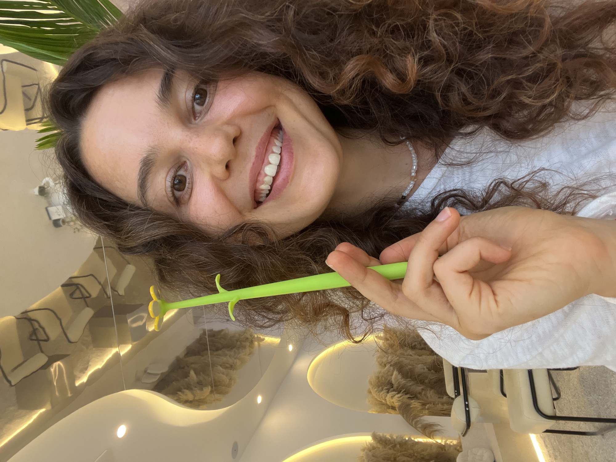 A young woman with curly brown hair smiles at the camera, holding a green flower-shaped pen. She wears a white shirt and a pearl necklace. The background features a modern interior with reflective ceilings, recessed lighting, and decorative dried plants. The scene is well-lit and appears to be indoors, possibly in a salon or boutique.