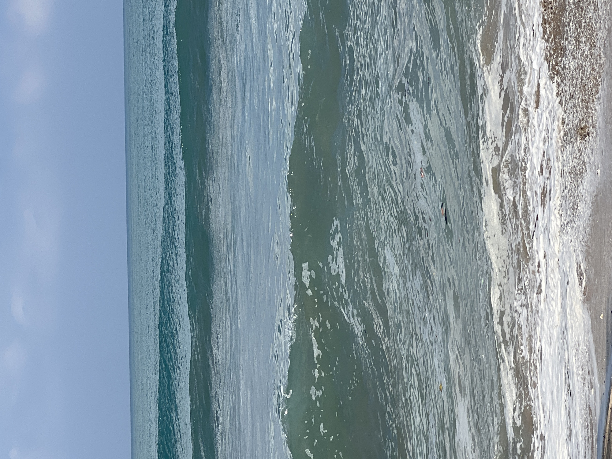 The image shows a view of the ocean with waves breaking on a sandy beach. The sky is clear and blue with some light clouds. The water is a deep blue-green color. There are several waves in the ocean, with the closest wave breaking and creating white foam. A few pieces of debris are visible floating in the water. The sand on the beach is wet and appears to be made up of small pebbles and shells. The scene is likely in Kuta, Indonesia, known for its beaches and surfing.