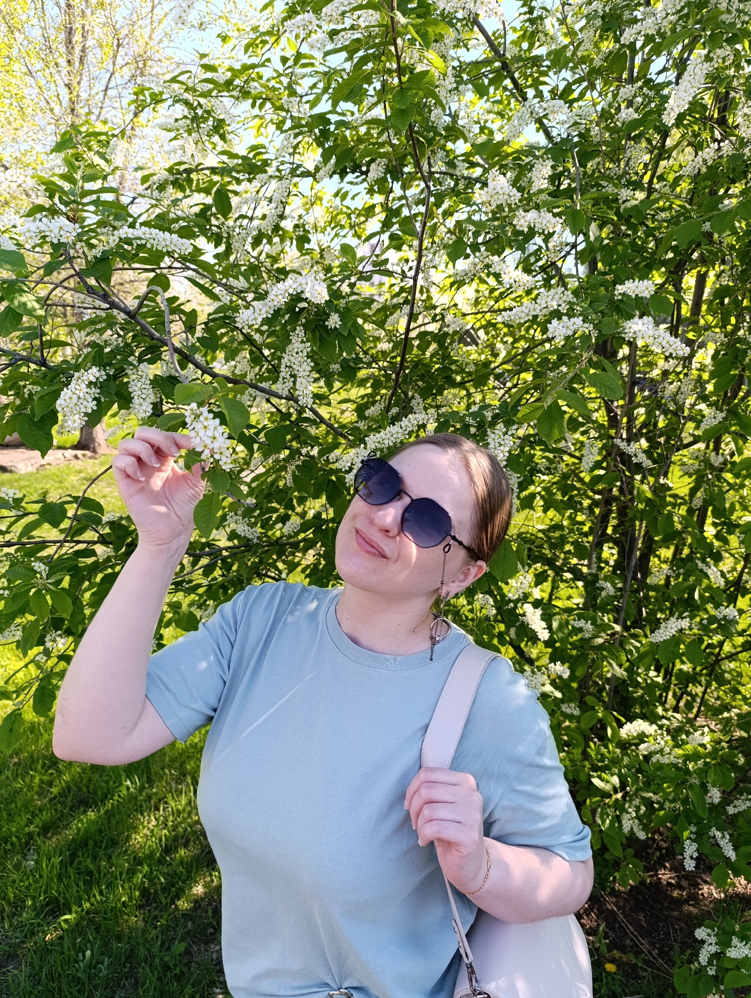The image depicts a woman standing in front of a flowering tree. She is wearing sunglasses, a light blue t-shirt, and a light-colored shoulder bag. She is holding a branch of white flowers with her right hand and gazing at them. The tree is in full bloom, with white flowers covering the branches. The background includes green grass and other trees. The location is a park or garden, but the specific city and location are unknown.