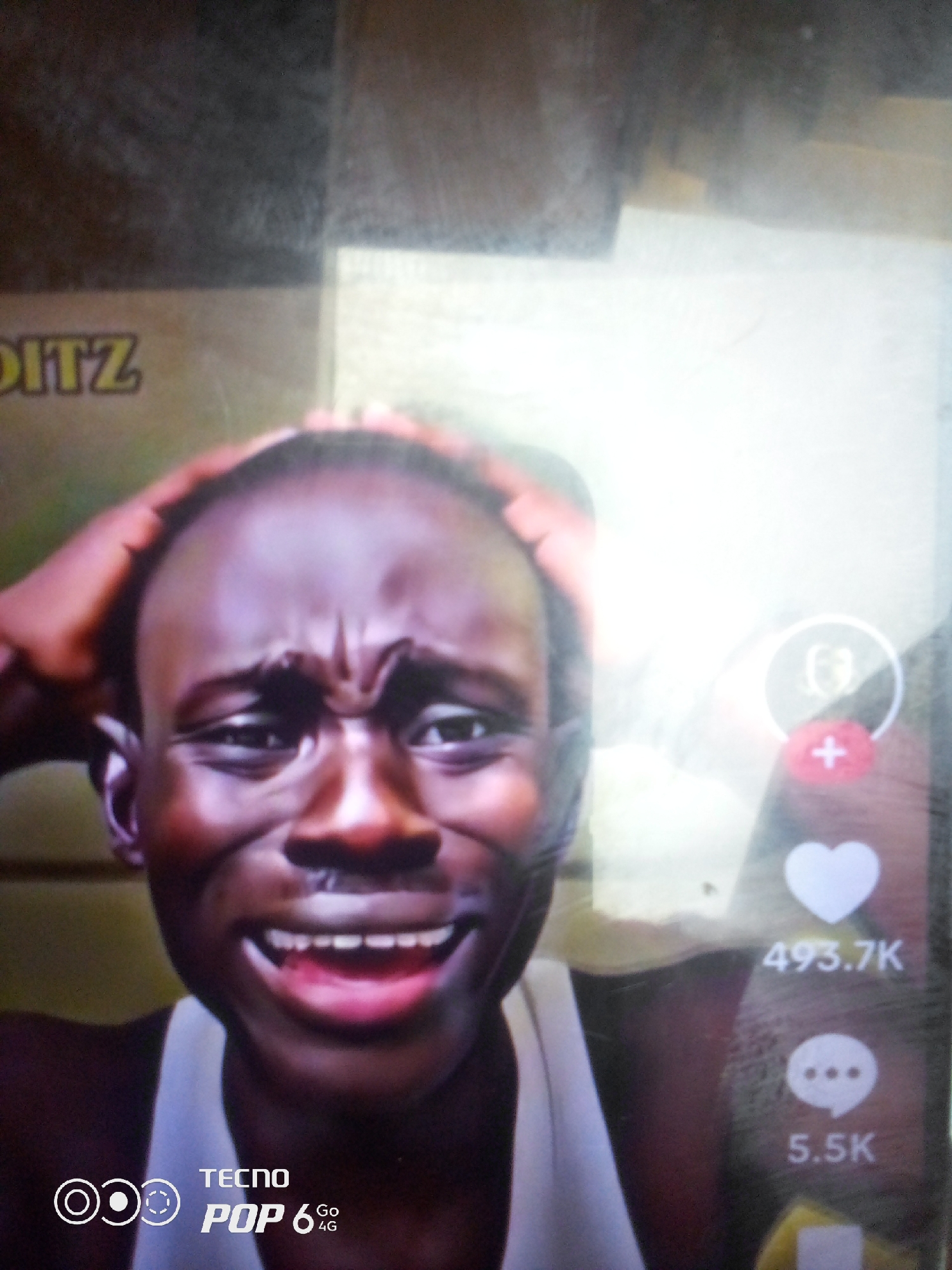 This is a close-up, low-angle, full-body shot of a Black male, seemingly in his late teens or early twenties, facing the camera. He has dark skin, a shaved head, and his hands are raised to his forehead, pressing down on his temples. His facial expression is contorted, with furrowed brows and an open mouth, revealing his teeth and pink gums, suggesting distress or extreme emotion. He is wearing a white tank top.
To the right of the subject, there are social media interface elements visible. A circular profile icon with a person inside is present, followed by a red circular icon with a white plus sign. Below that, a white heart icon is displayed with the number "493.7K" beneath it, indicating likes. Further down, a speech bubble icon with three dots is shown, with "5.5K" below it, likely representing comments.
In the bottom left corner, a logo for "TECNO POP 6 Go 4G" is visible, with accompanying circular graphic elements.
The background is indistinct, appearing to be a mixture of dark and brightly lit areas, possibly indicating a screen or an environment with artificial lighting. The overall image quality is somewhat grainy and appears to be taken from a digital device, possibly a phone.
There is no indication of the specific location in Howayi, Nigeria, within the provided image.
