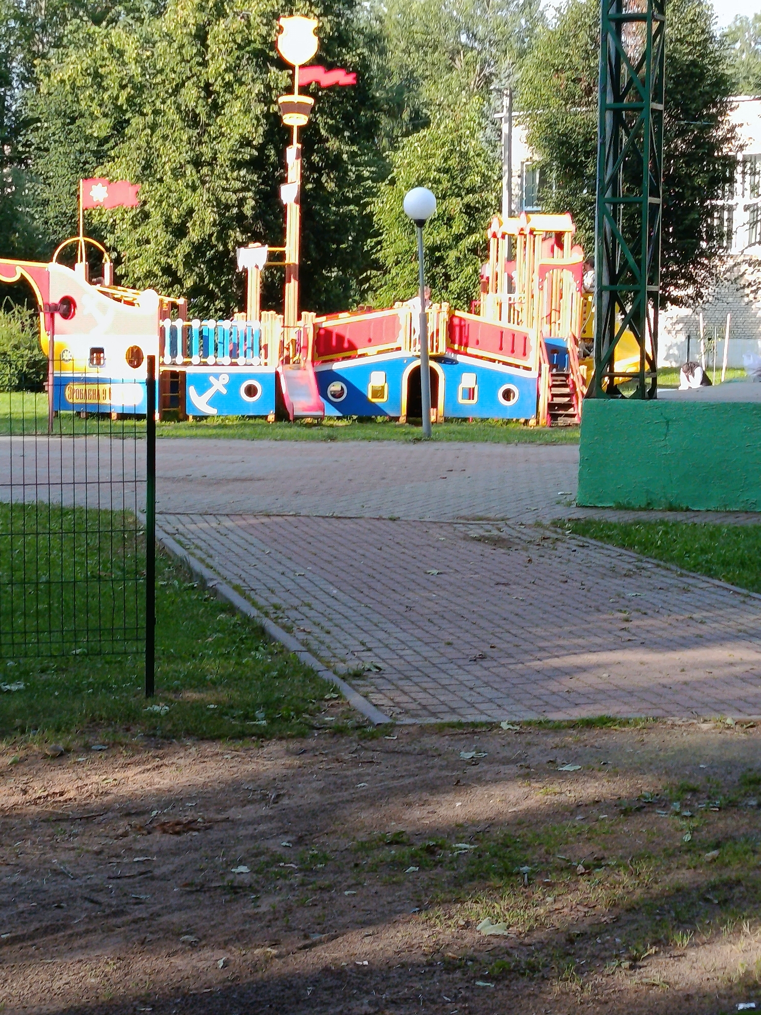 The image captures a children's pirate ship playground on a sunny day, situated in an outdoor area. The playground structure is colorful, featuring red, blue, and yellow elements, with a prominent mast topped by a golden emblem and red flags. An anchor symbol is visible on the blue hull. The playground is surrounded by greenery, including trees and grass. A paved walkway made of interlocking bricks runs in front of the playground, and a wire fence encloses part of the grassy area. In the background, a building with windows can be seen, and a tall green metal structure is present to the right of the playground. A street lamp with a spherical white light is also visible. The scene appears to be a public park or recreational area in the city of Smilyavichy, within the Smilyavichy rural council, Belarus.