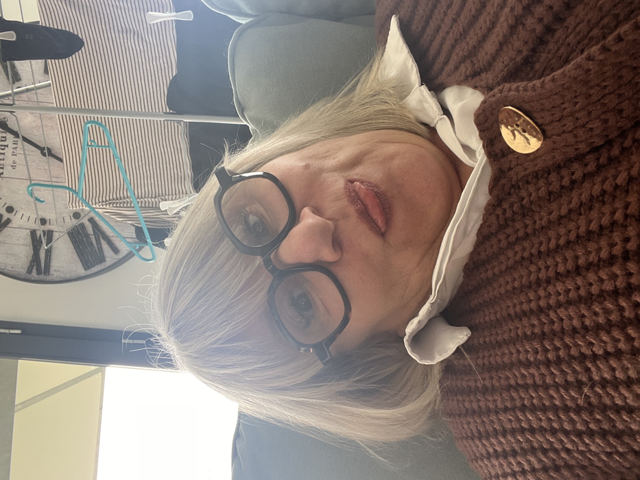 This close-up image depicts a fair-skinned individual, likely female, with shoulder-length blonde hair and bangs, posing for a selfie indoors in Chambœuf, France. The person wears dark-framed eyeglasses and has their tongue slightly extended from their mouth, indicating a playful or informal expression. They are wearing a white collared shirt underneath a brown, coarsely knit cardigan, fastened with a single, large, irregular gold-colored button on the right side of the chest. The individual's head is tilted slightly to the left, and they are situated against a light green cushion, likely part of a sofa.
In the background, visible to the upper right of the individual's head, is a large, round, distressed-style wall clock with Roman numerals, featuring text that appears to read "ANTIQUES de PARIS." Below and in front of the clock, a light blue plastic clothing hanger is visible on what appears to be a metal clothes drying rack. Various garments, including a light-colored striped shirt and dark clothing items, are hanging on the rack, secured by white plastic clothespins. To the upper left, a bright, possibly overcast sky or an overexposed window is visible.