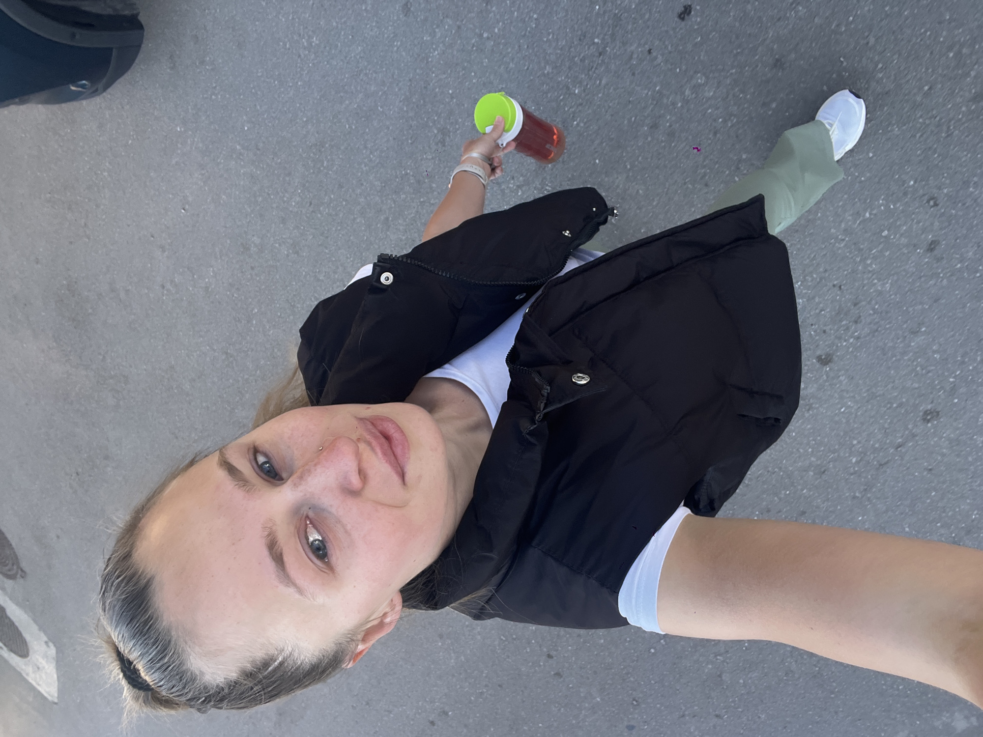 A person is outdoors on a paved surface, likely a street, based on the gray color and texture of the ground. The perspective is from a high angle, suggesting the photo was taken from above or by the person themselves. The person is wearing a black vest over a white top, light green pants, and white sneakers. They hold a container with a green lid in their left hand. The background is nondescript, showing only the pavement and a portion of a dark vehicle in the upper left corner. Specific details regarding Bečići, Montenegro are not discernible from this image.