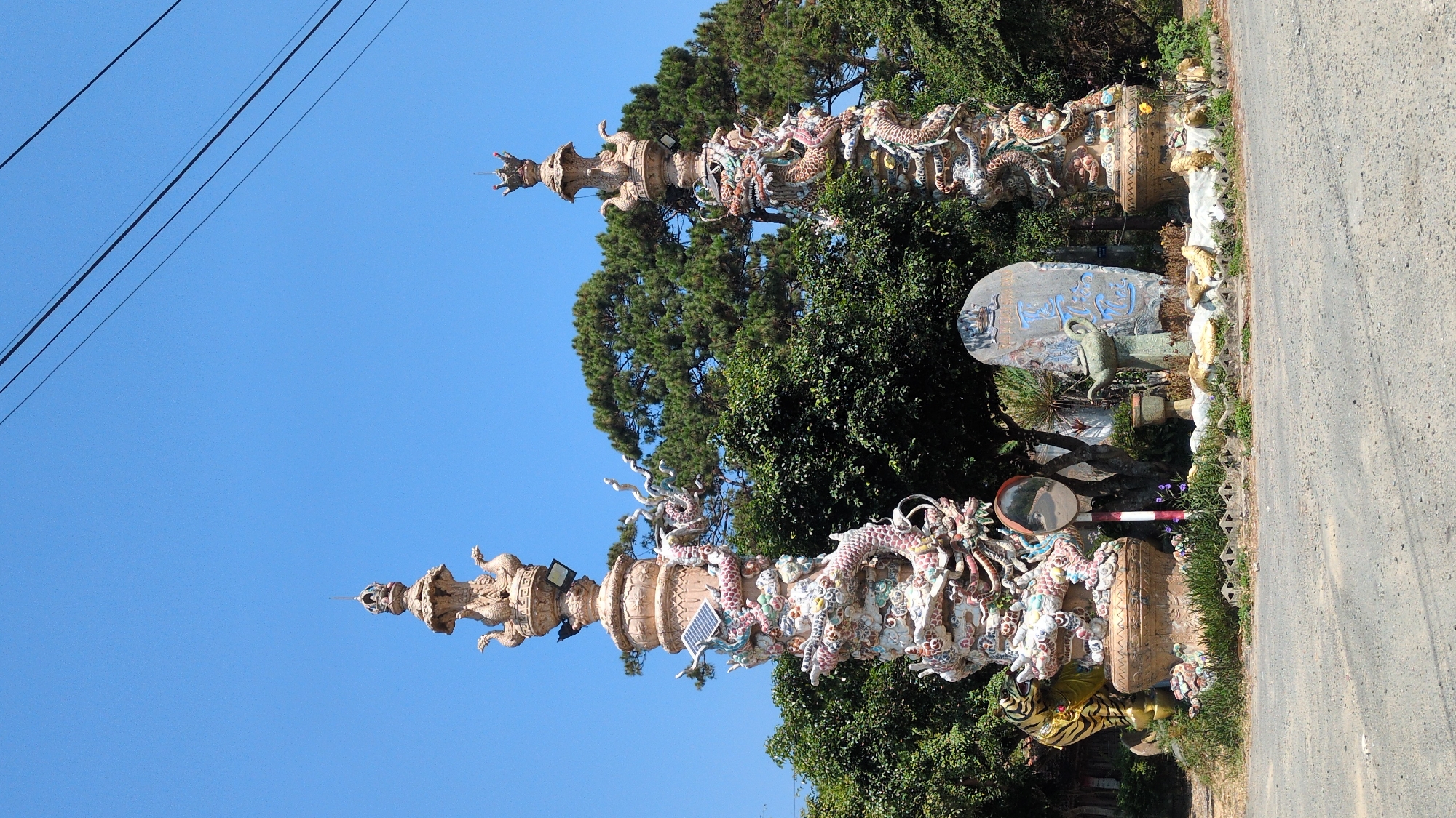 The image shows two ornate, dragon-covered pillars located in Lâm Sơn, Vietnam. The pillars are made of a light-colored material and are adorned with colorful ceramic dragons. They are positioned alongside a road and surrounded by lush greenery and trees. The sky is clear and blue, with power lines visible in the upper left corner. A weathered sign with blue characters is visible between the pillars. Various objects such as a mirror, a pot, and what appears to be a solar panel are incorporated into the display.