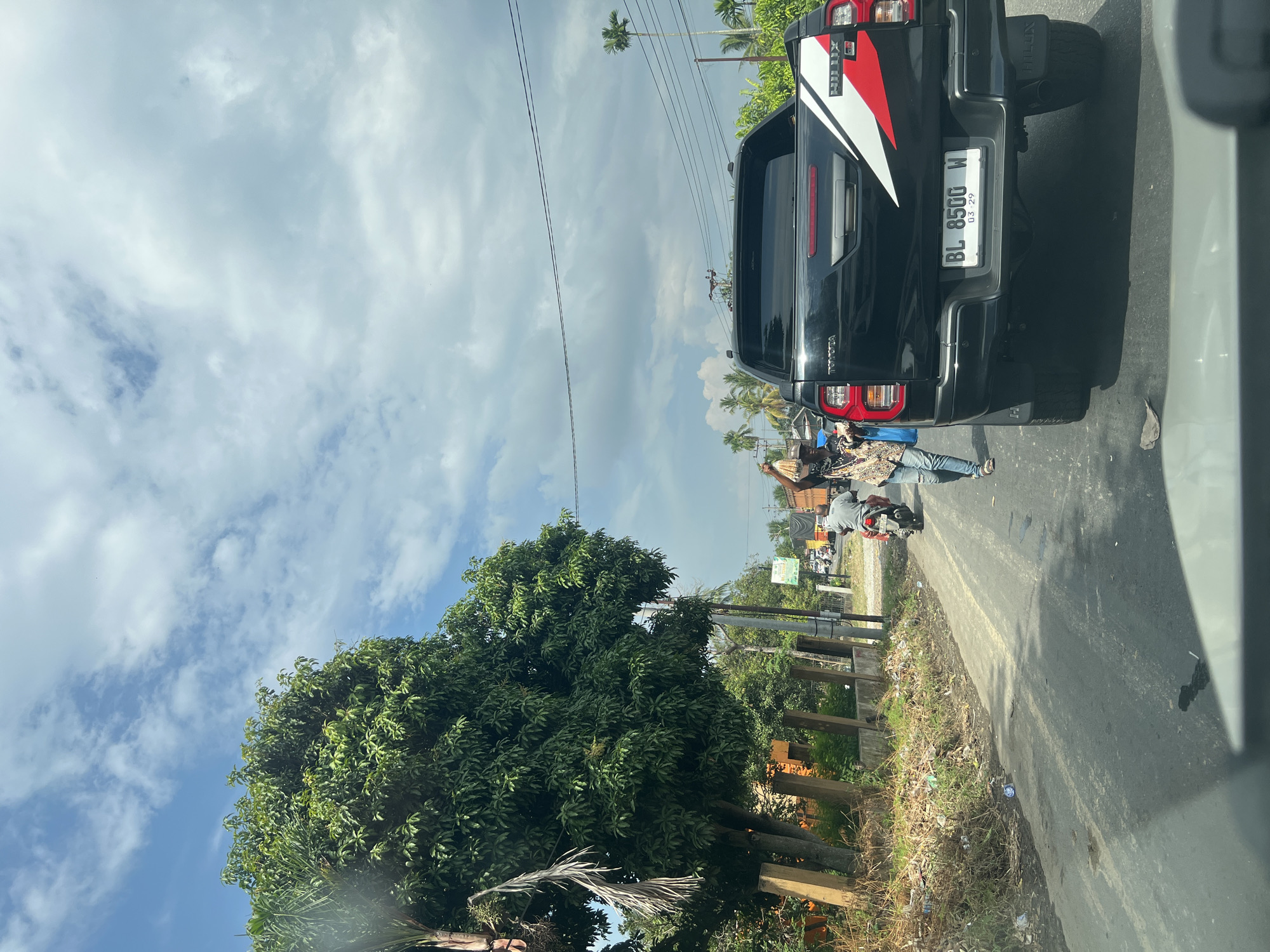 The image depicts a street scene in Blangpanjoe, Indonesia. A black Toyota Hilux pickup truck is prominently featured in the foreground, with its license plate reading "BL 8500 W." To the left of the truck, a person is walking along the side of the road, carrying bags. A motorcycle is also visible on the road, with a rider. In the background, there is a large tree with dense foliage, along with other vegetation and buildings. The sky is partly cloudy, with a mix of blue sky and white clouds. The scene appears to be a roadside area with some commercial activity.