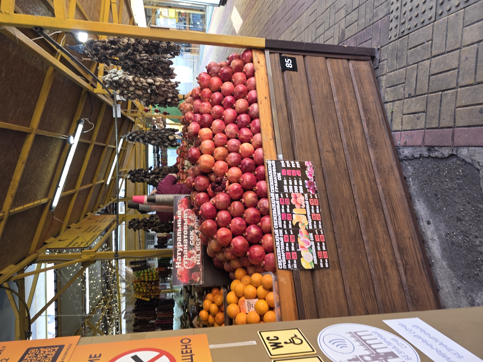 This image depicts a bustling market stall in Sochi, Russia, likely during the daytime. The stall features a yellow metal frame supporting a wooden structure, identified by the number "85."
The main attraction is a large display of fresh, red pomegranates meticulously stacked in a pyramid shape. Below them, several crates are filled with bright orange tangerines or small oranges. Hanging prominently from the stall's overhead structure are numerous bundles of dried produce, possibly dried fruits, vegetables, or spices.
A person wearing a purple top is partially visible behind the fruit display, presumably working at the stall.
Several signs in Russian detail the offerings. A large sign prominently advertises "Натуральный гранатовый СОК на все 100%" (Natural pomegranate juice 100%). Another sign presents a price list for "СВЕЖЕВЫЖАТЫЙ" (Freshly squeezed) juices, including "ГРЕЙПФРУТОВЫЙ СОК" (Grapefruit juice), "АПЕЛЬСИНОВЫЙ СОК" (Orange juice), and "ГРАНАТОВЫЙ СОК" (Pomegranate juice), with prices listed in rubles for various volumes (e.g., 100 ml, 200 ml, 500 ml). Other visible text includes "WC" with an accessibility symbol, and "БЕЗНАЛИЧНЫЙ" (Cashless payment). The partial text "ADLERRYWOKZ" likely transliterates to "Adlerrynok," indicating the market is in the Adler district of Sochi.
The ground is paved with rectangular and square bricks in shades of grey and brown, characteristic of an outdoor or semi-enclosed market environment. The scene conveys a sense of a lively, fresh produce and juice vendor.