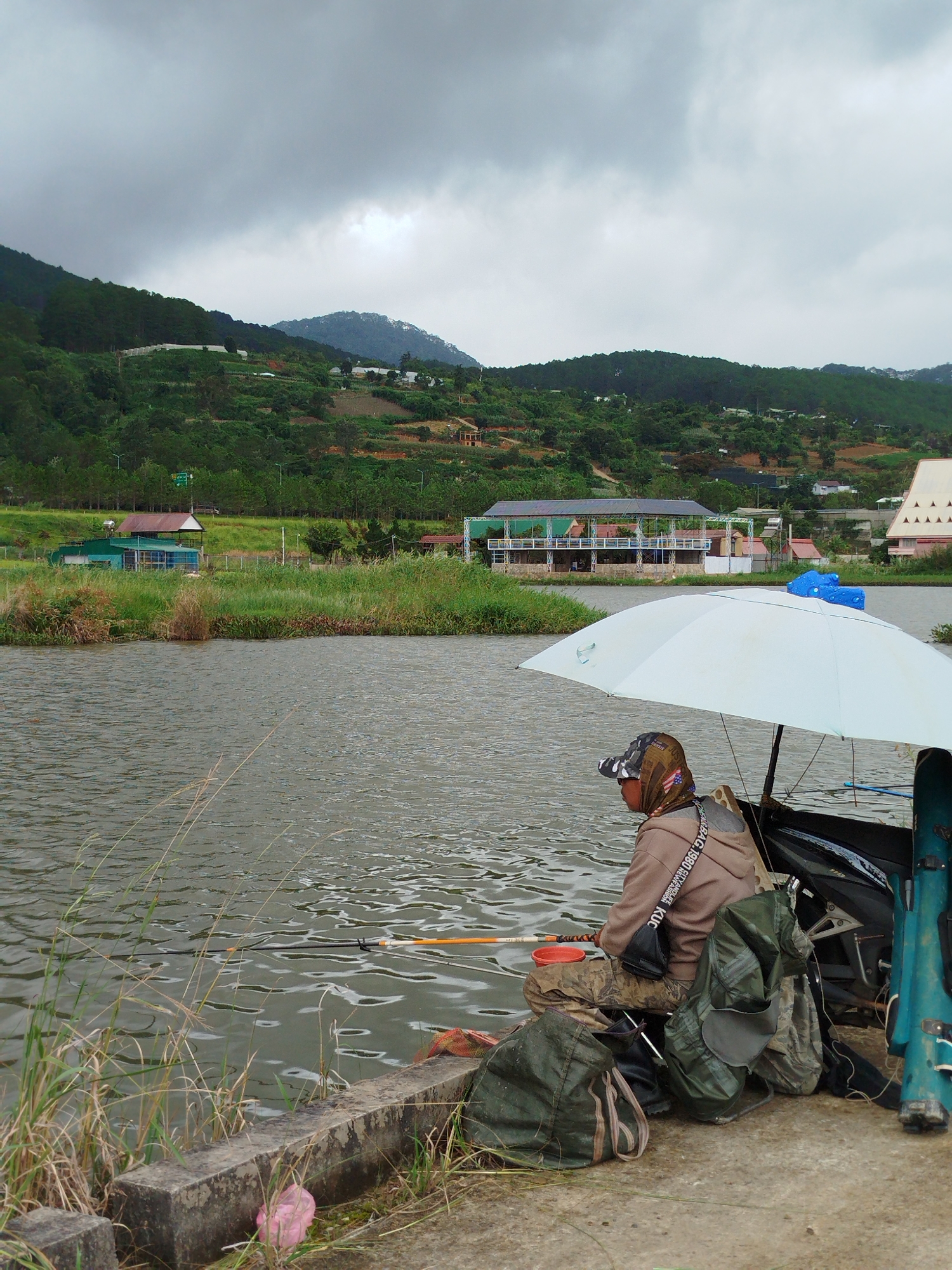 The image depicts a person fishing from a concrete bank overlooking a body of water. The individual is seated, wearing camouflage pants and a brown hooded sweatshirt, with a patterned bandana covering their head. They are positioned under a large, light-colored umbrella, with fishing rods extended into the water. Several bags, including a green backpack and a dark green duffel bag, are situated around the person. In the background, across the water, is a lush green hillside dotted with buildings and agricultural terraces, characteristic of the landscape around K'Long, Vietnam. The sky is overcast, suggesting a cloudy or possibly rainy day. The scene conveys a sense of quiet contemplation and outdoor recreation.