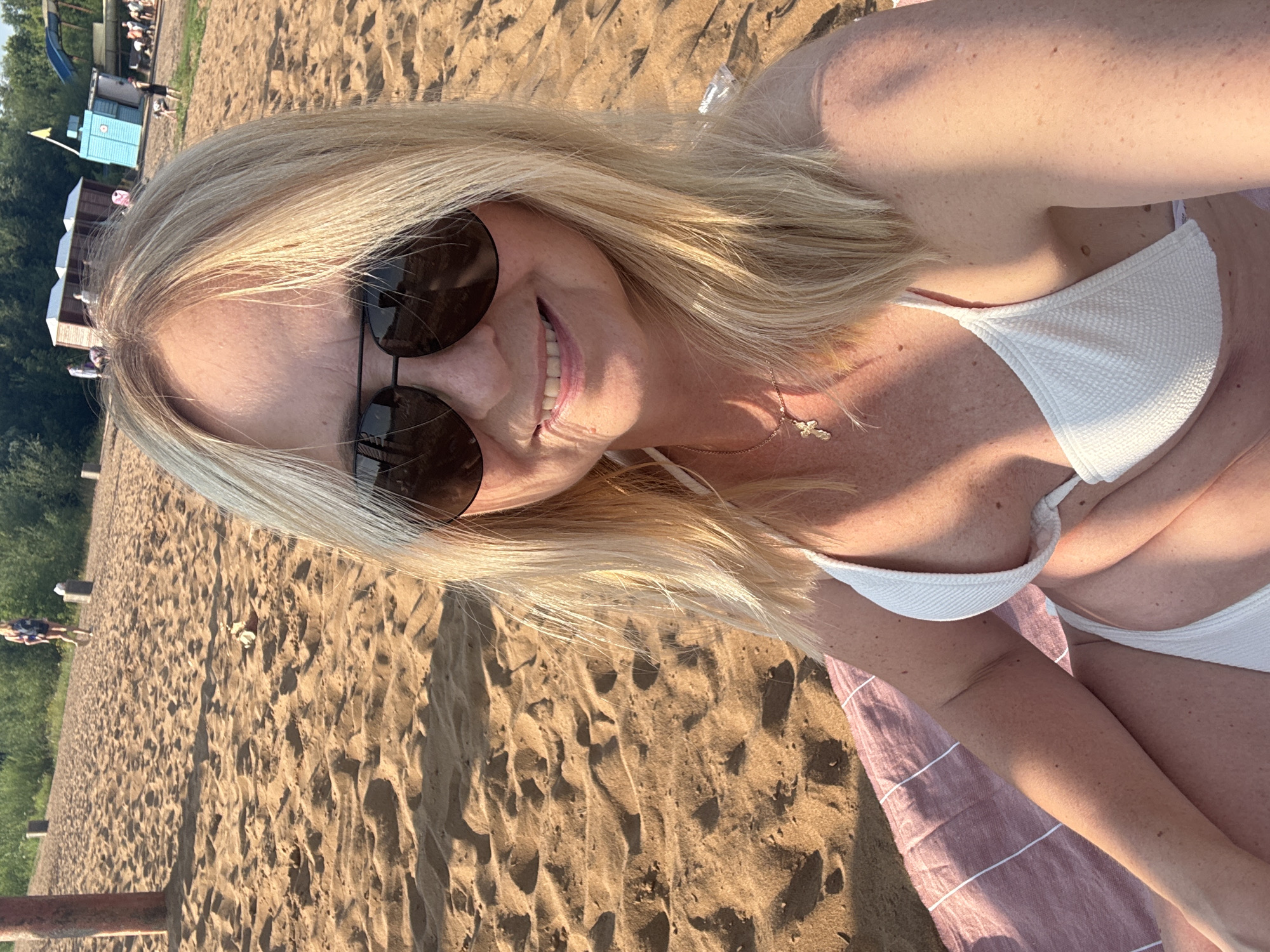 A woman is sitting on a beach, taking a selfie. She has blonde hair, sunglasses, and is wearing a white bikini. The beach has sand and is next to a body of water. There are some trees and buildings in the background. The sun is shining, casting shadows on the sand.