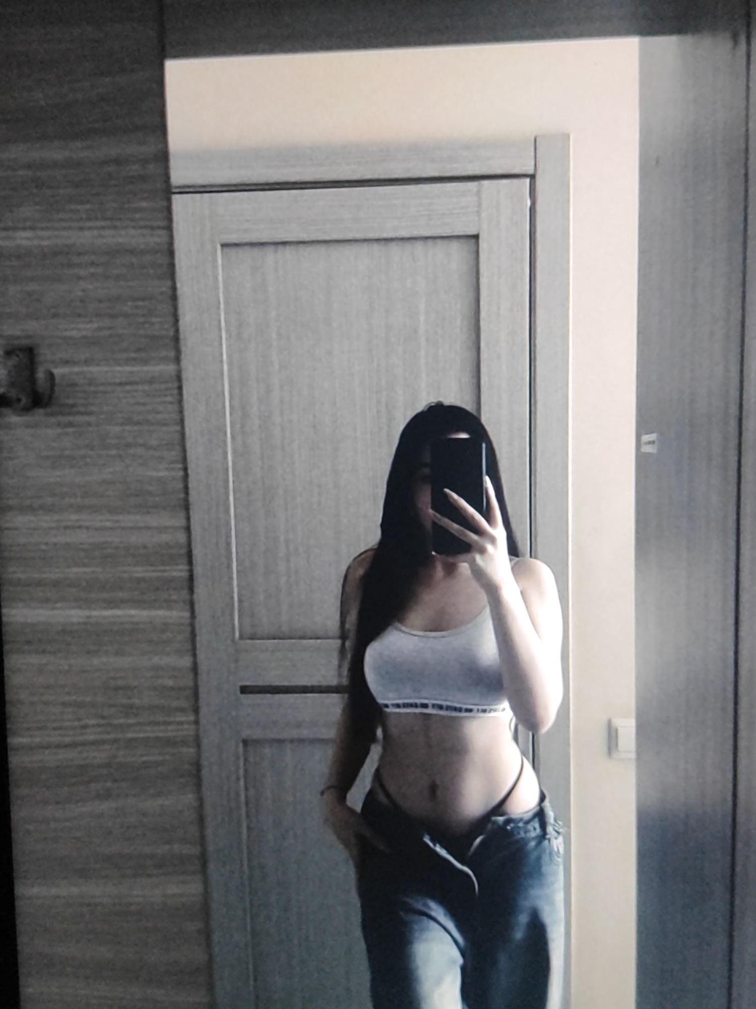 A person is taking a mirror selfie in what appears to be a room with a door and a wall. The person is wearing a crop top and jeans. The lighting is dim, and the image quality is low. The context of Fergana City, Uzbekistan, is not evident from the image itself.