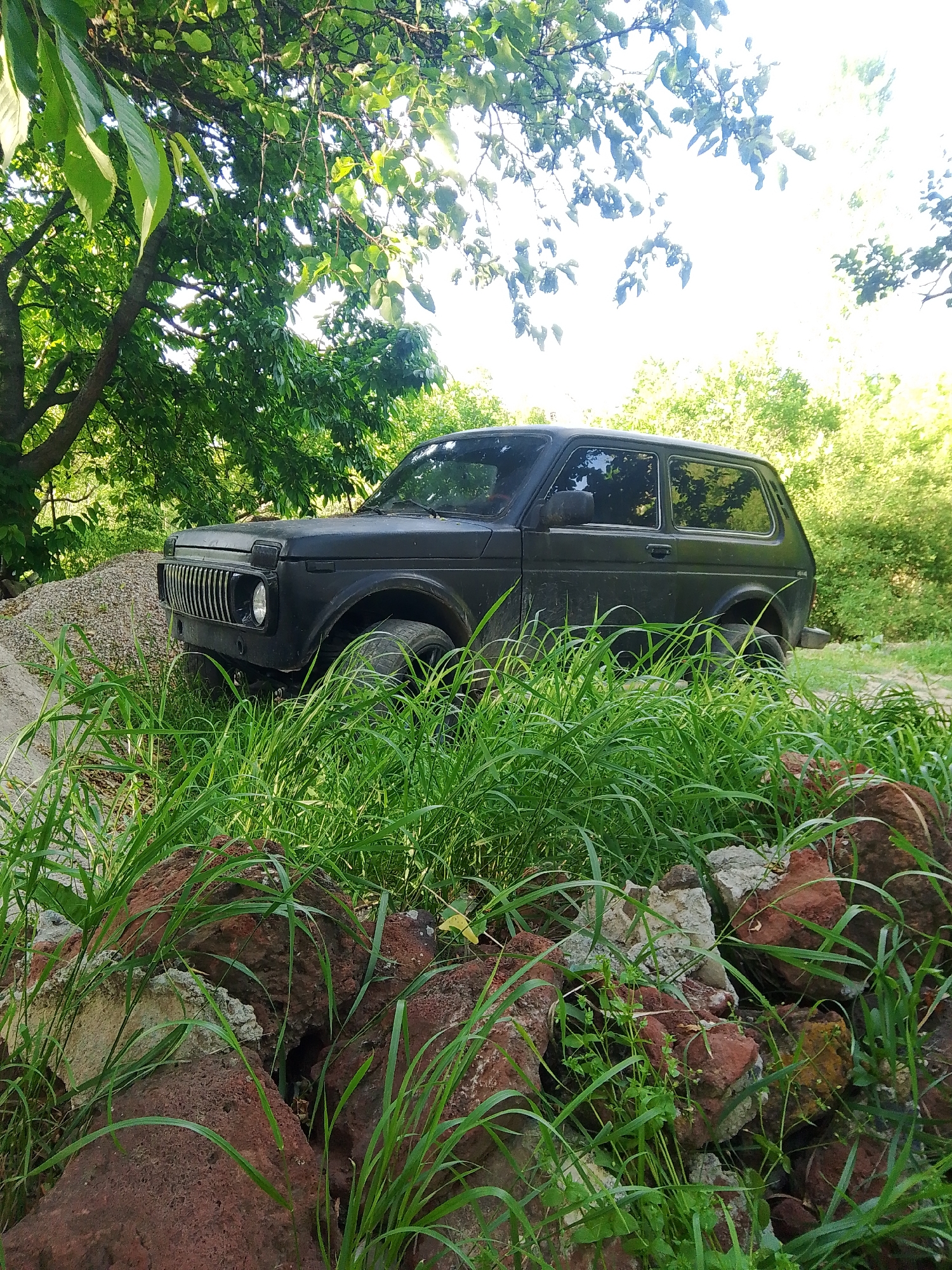 A black three-door Lada Niva SUV is positioned on an unpaved, uneven surface in Parpi, Armenia. The vehicle's front is angled towards the left, displaying its distinctive vertical slat grille and a single circular headlight on the passenger side. The body of the vehicle appears matte black and slightly dirty or weathered. To the immediate left of the car's front bumper, a substantial pile of light-colored gravel or crushed stone is visible.
The foreground is characterized by dense, vibrant green tall grass that partially obscures the vehicle's lower half. Among the grass, several reddish-brown, porous rocks or pieces of broken concrete debris are scattered.
In the mid-ground and background, natural vegetation dominates. A large tree with a thick, dark trunk and abundant green foliage occupies the upper left portion of the image. Sunlight filters intensely through the tree canopy, creating bright highlights on the leaves. The upper right section of the image shows an intensely bright, possibly overexposed sky, indicating strong sunlight. Further to the right in the background, a dense cluster of green trees and foliage is present. The overall environment appears rural or semi-rural, lacking overt signs of urban infrastructure. No individuals are present in the scene, and no actions or interactions are depicted. The scene is static and brightly illuminated by natural daylight.