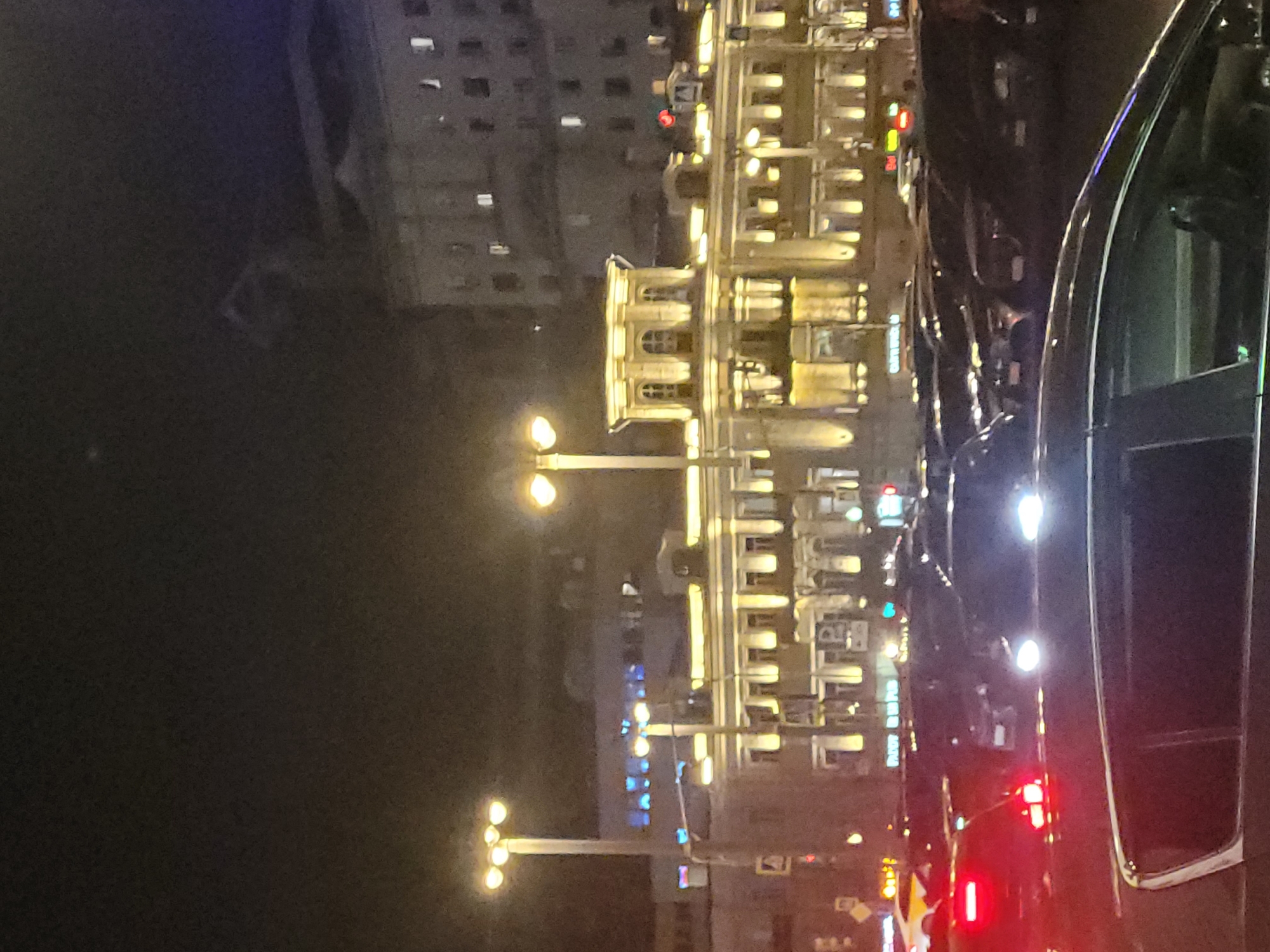 The image depicts a nighttime scene in Moscow, Russia. A multi-story building illuminated with yellow lights lines the street. Streetlights with multiple bulbs extend over the road. Cars are lined up on the street, headlights creating bright spots in the darkness. The side of a dark car is visible in the foreground. Other buildings can be seen in the background.