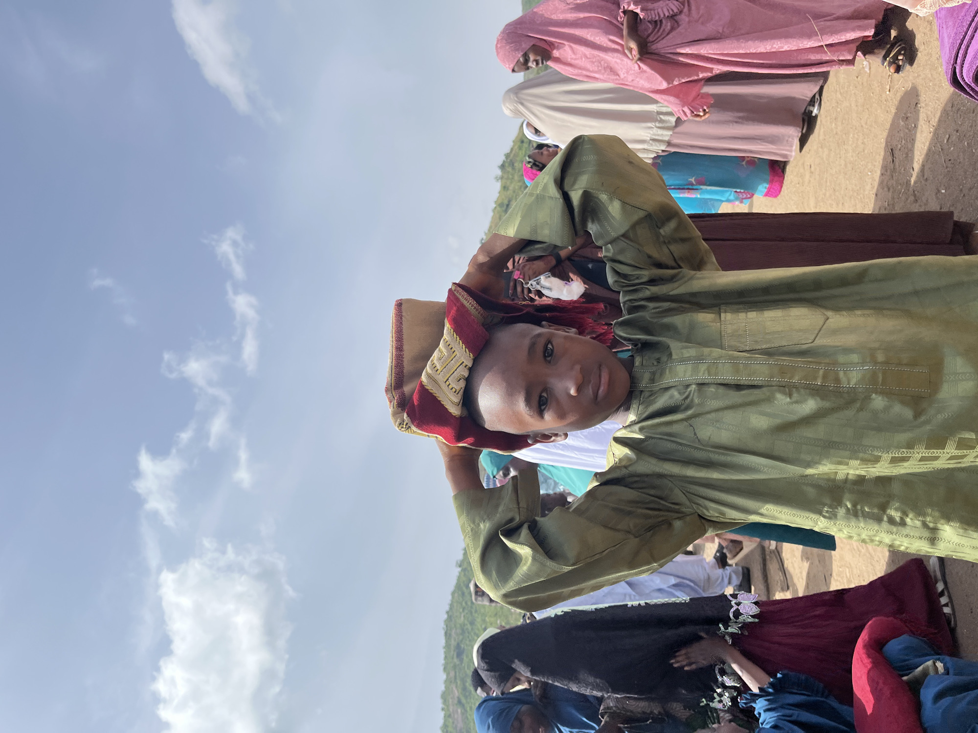 A boy wearing a green garment and a colorful hat stands in a gathering of people in Naraguta Hausawa, Nigeria. The sky is visible in the background. The boy holds his hands up to his hat. The people are wearing diverse colored clothing.
