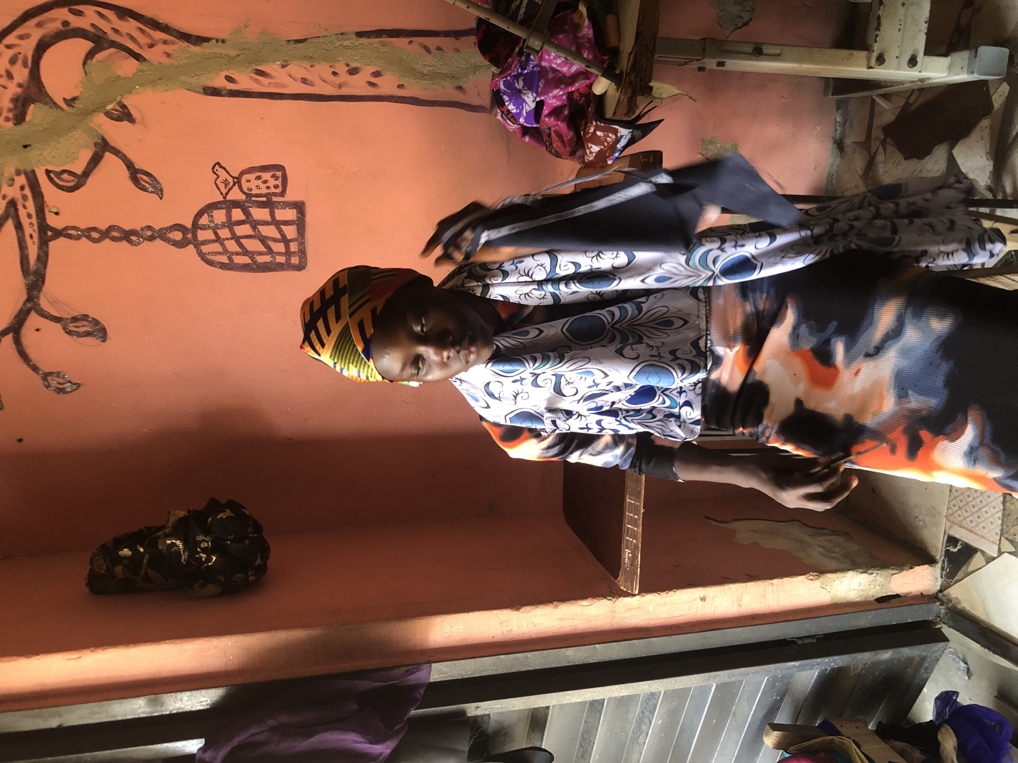 A young girl is sitting in what appears to be a workshop in Ado, Nigeria. She is wearing a colorful headwrap and a matching outfit with intricate patterns. In her right hand, she is holding a dark fabric. The wall behind her is painted a pale orange and decorated with a drawing of a tree with a birdcage hanging from a branch. To her left, there is a shelf with some fabrics and to her right, a sewing machine. The room is lit by natural light, casting shadows on the floor.