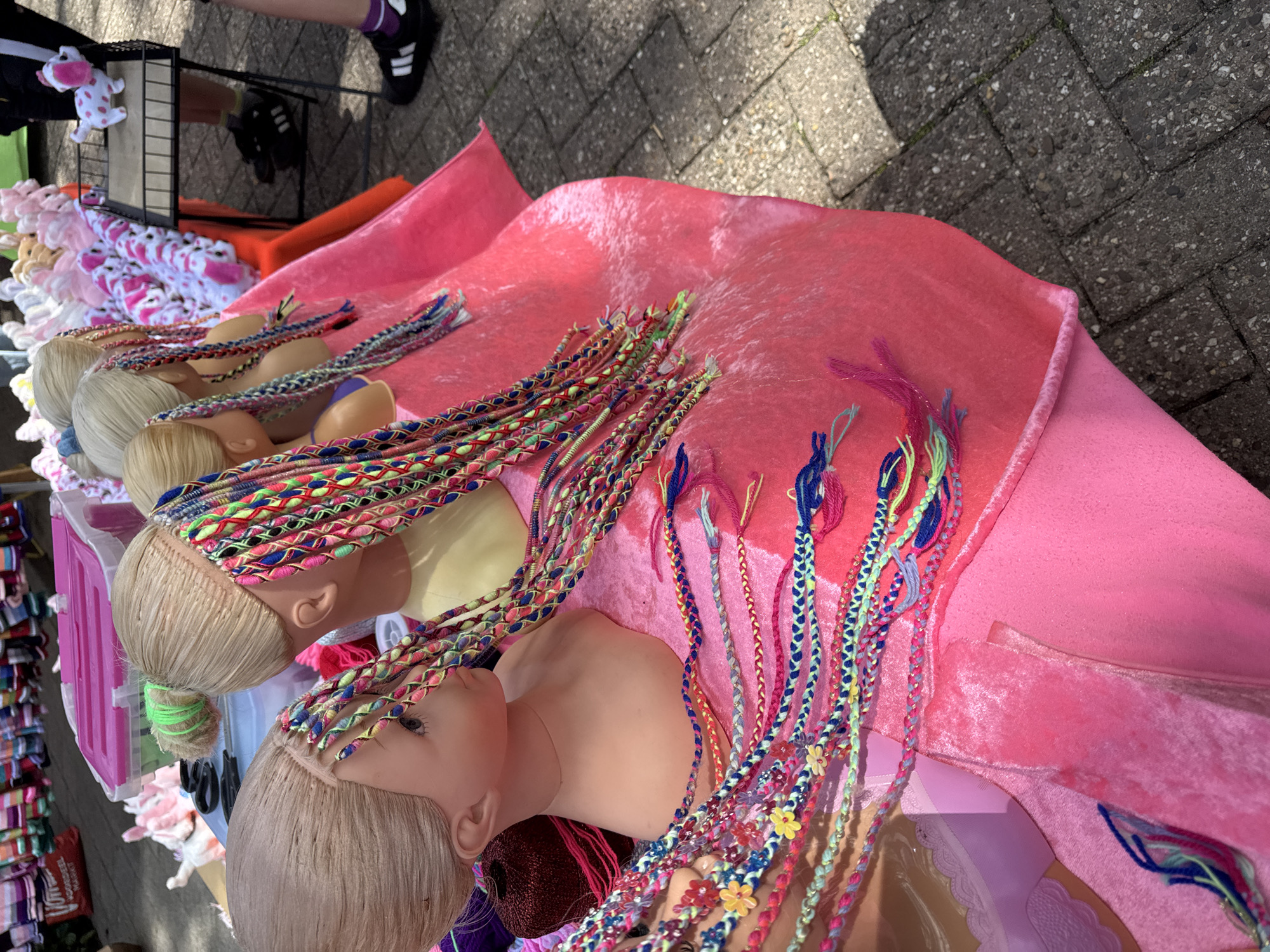 An outdoor market stall displays several mannequin heads adorned with colorful braided hair extensions. The heads are positioned on a pink velvet-covered surface. In the background, a pink plastic bin and racks filled with stuffed animals are visible. A person's legs, wearing black sneakers and purple socks, are also visible on the right side of the image. The ground is paved with stone tiles.