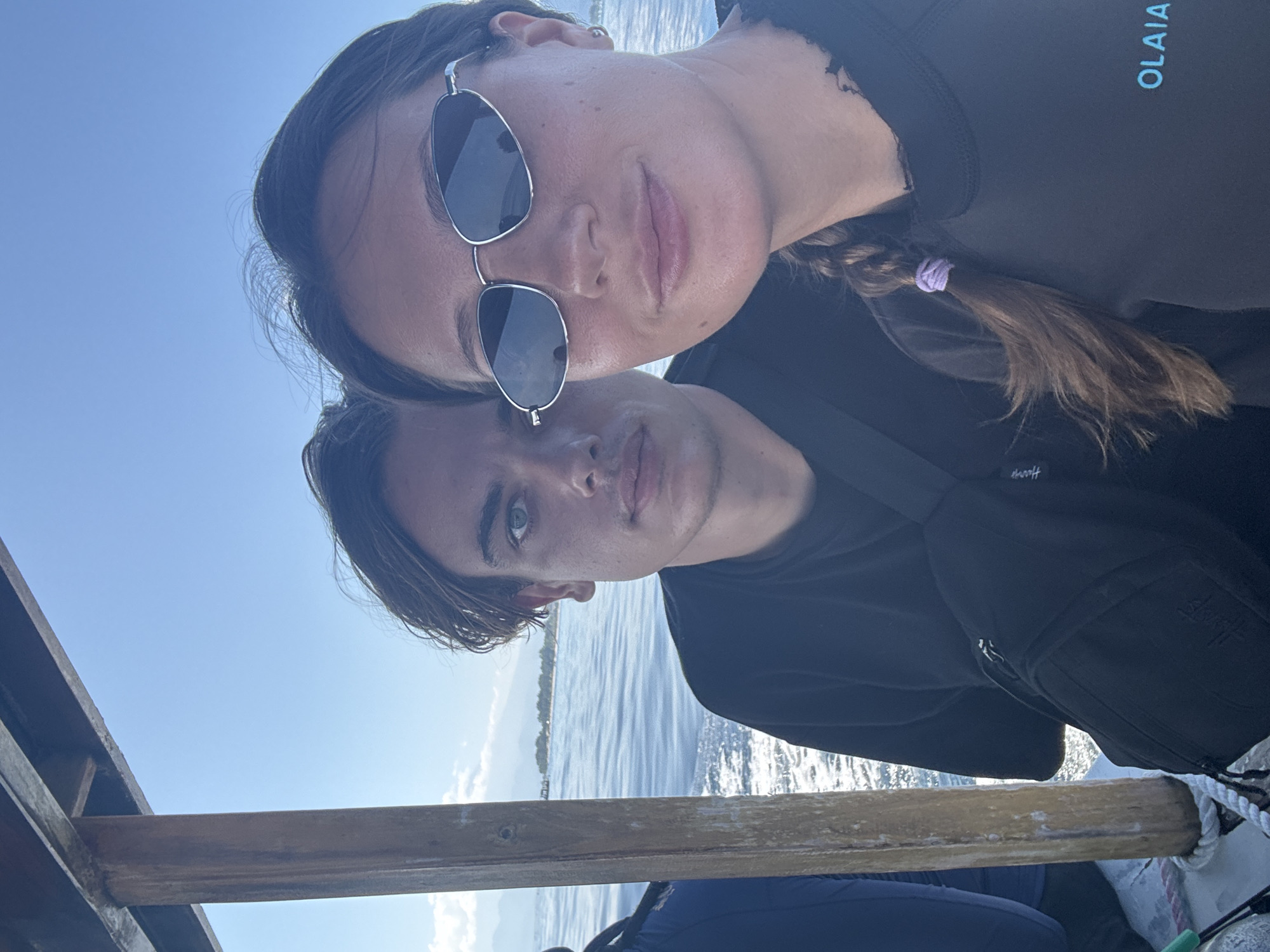 A selfie depicts two individuals in an outdoor setting, likely on a boat. A woman with sunglasses and a braid is positioned in front, while a man is behind her. The background reveals a body of water and faint landscape. The location is Unknown, Indonesia.