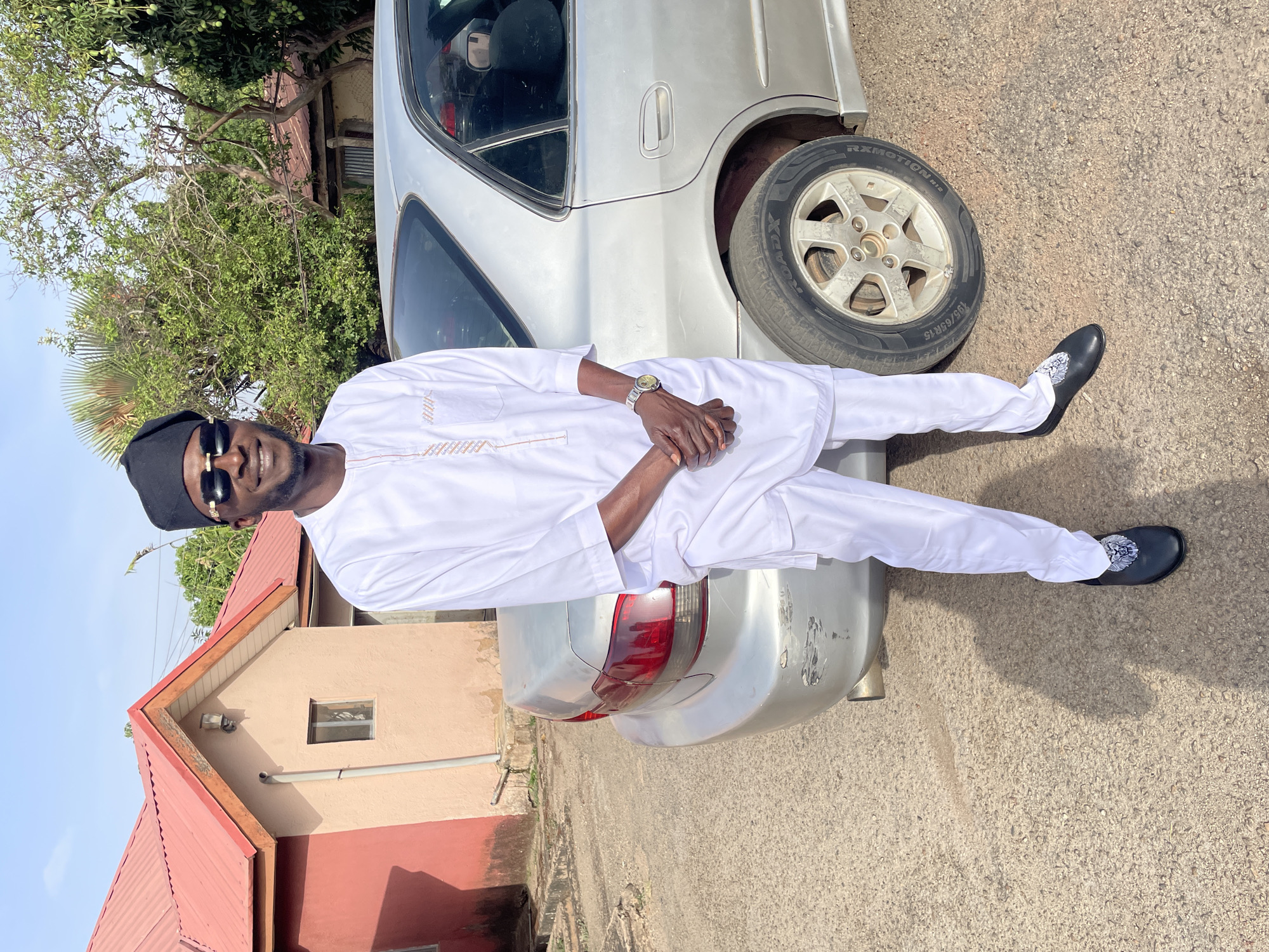 The image depicts a man posing on a silver car. He is wearing a white garment, a black cap, and sunglasses. The location is an outdoor environment next to a building with a red roof. The city and country are unknown. The man is leaning on the car with his arms crossed and his legs outstretched.