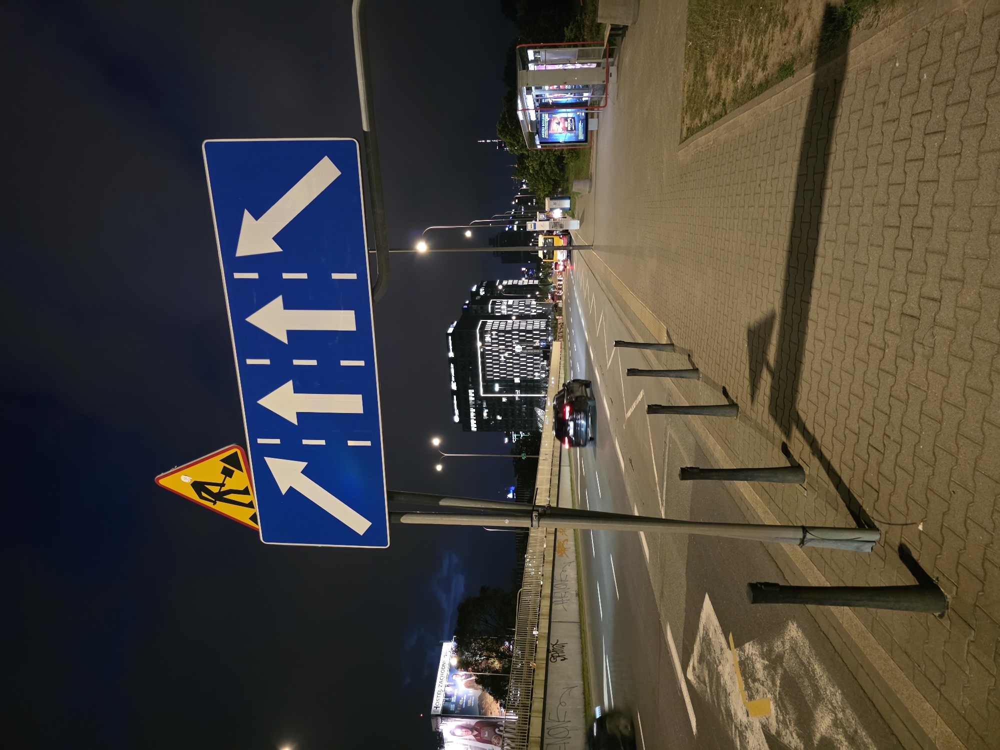 The image captures a night scene in Warsaw, Poland, focusing on a street with traffic signs and urban infrastructure. Prominently displayed is a blue rectangular traffic sign with white arrows indicating multiple lanes, with one lane diverging to the left. Above this sign, a yellow triangular warning sign depicts a construction worker with a shovel. The street is illuminated by streetlights and the lights from surrounding buildings. A dark-colored car is visible on the road, and a bus and a bus stop are situated on the sidewalk to the right. The sidewalk is paved with interlocking concrete blocks, and metal bollards line the edge of the road. In the background, modern buildings with illuminated windows are visible, along with billboards and trees. The sky is dark, suggesting it is nighttime.