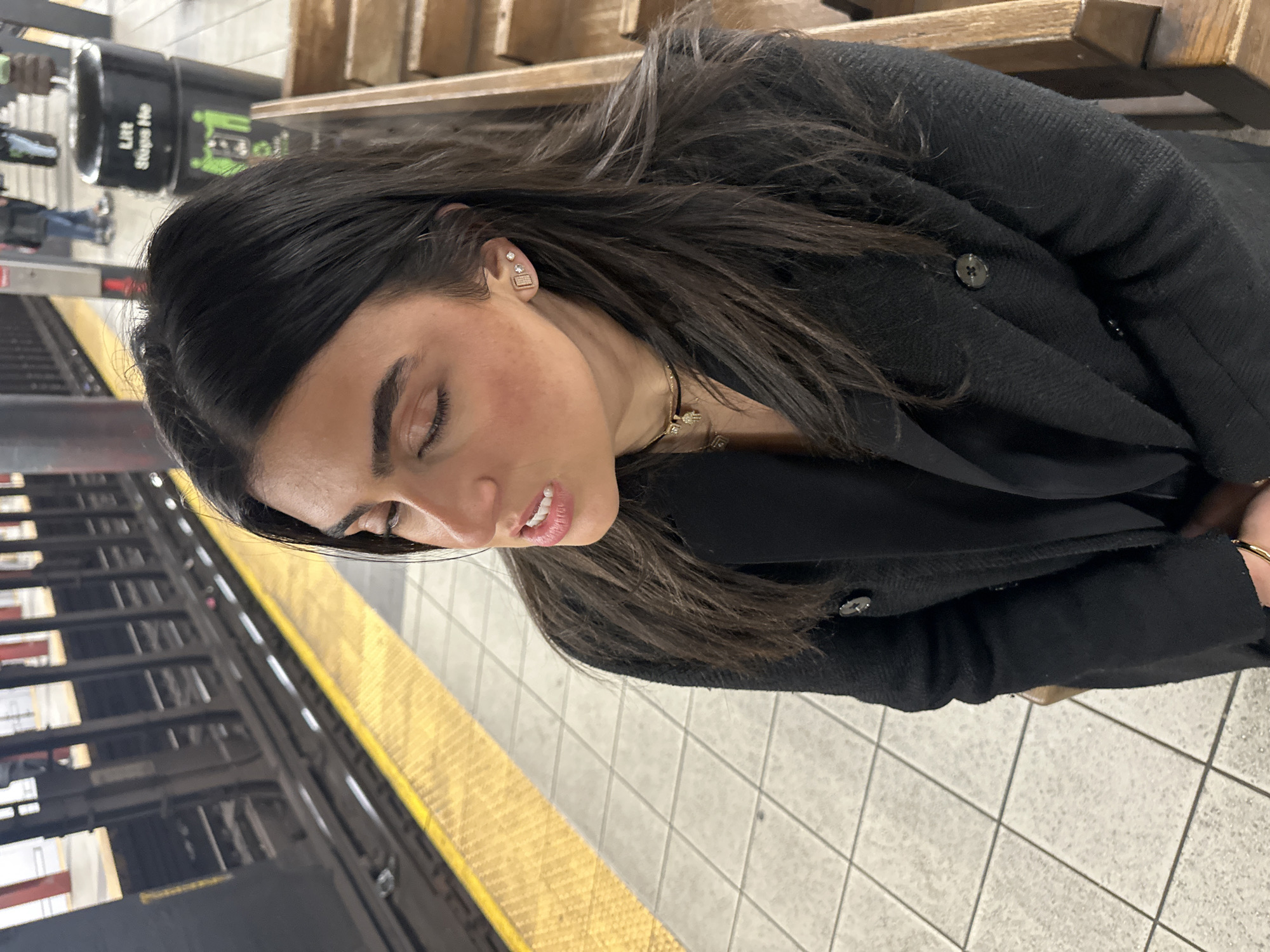 A woman is depicted seated on a subway platform in New York City, United States of America. She has long, dark hair and wears a black, textured coat over a dark top. Her eyes are closed or directed downward, and her mouth is slightly open, revealing her upper teeth. She wears a gold-colored bracelet on her left wrist, multiple necklaces (including a choker with a charm and a longer chain with a pendant), and three earrings on her visible right ear (a rectangular stud, a small circular stud, and another tiny stud). To her left, subway tracks are visible behind a yellow safety strip, with a train structure partially obscured. To her right and in the background, sections of a wooden bench are visible, along with a black trash bin labeled "Litt Stops He" and blurry figures of other people on the platform. The platform floor is composed of light grey square tiles, indicative of an underground subway environment.