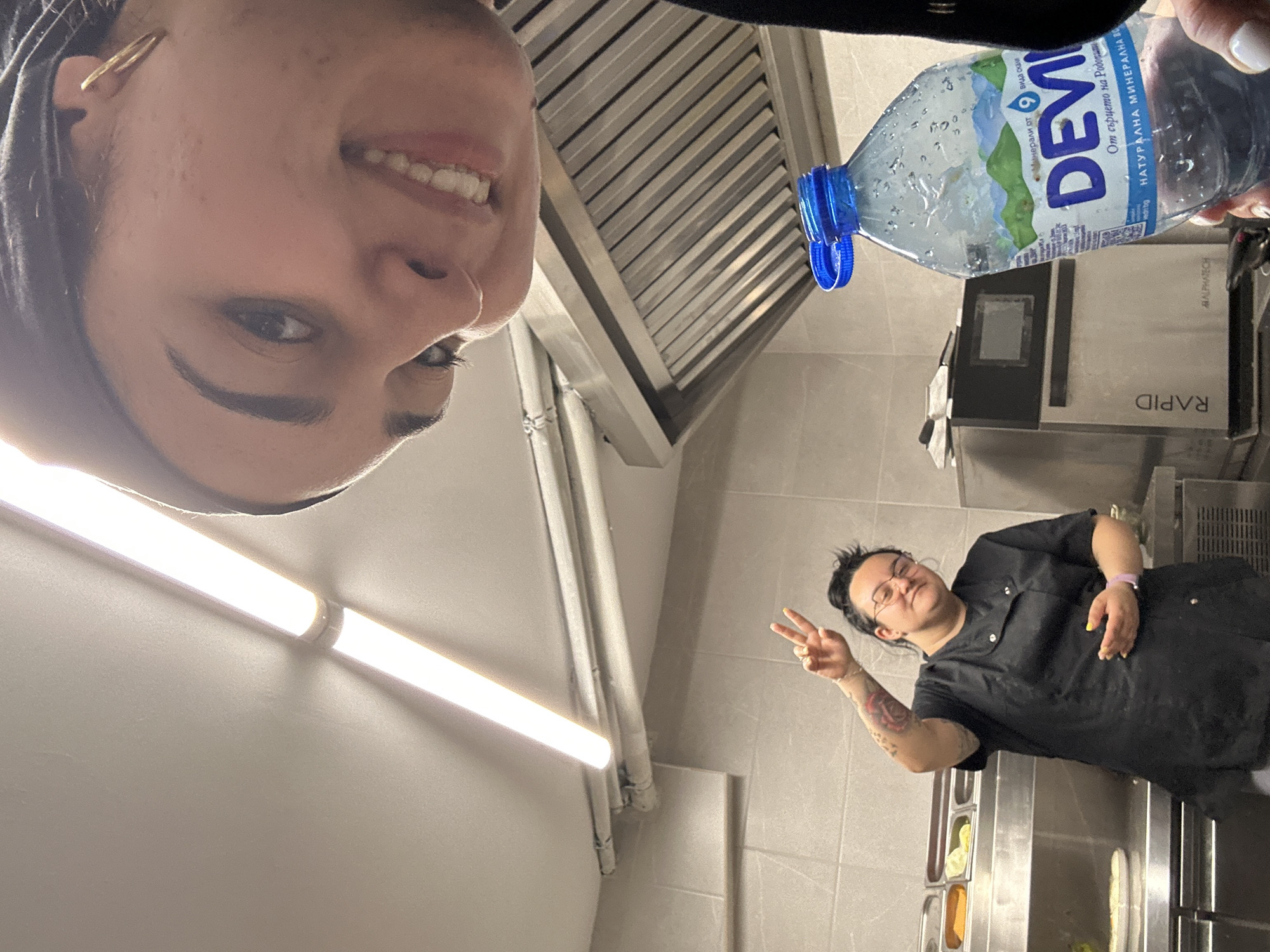 This is an upside-down selfie taken by a person showing part of their face, including their smile, eye, and ear with an earring. Another person in a black uniform is further away, giving a peace sign. They are standing in what appears to be a kitchen setting. In the background are kitchen appliances, including a "RAPID" machine and an exhaust hood. A water bottle labeled "DEVI" is also visible, with text that translates to "from the heart of Rhodopes." The location in Burgas, Bulgaria, cannot be determined from the provided image details.