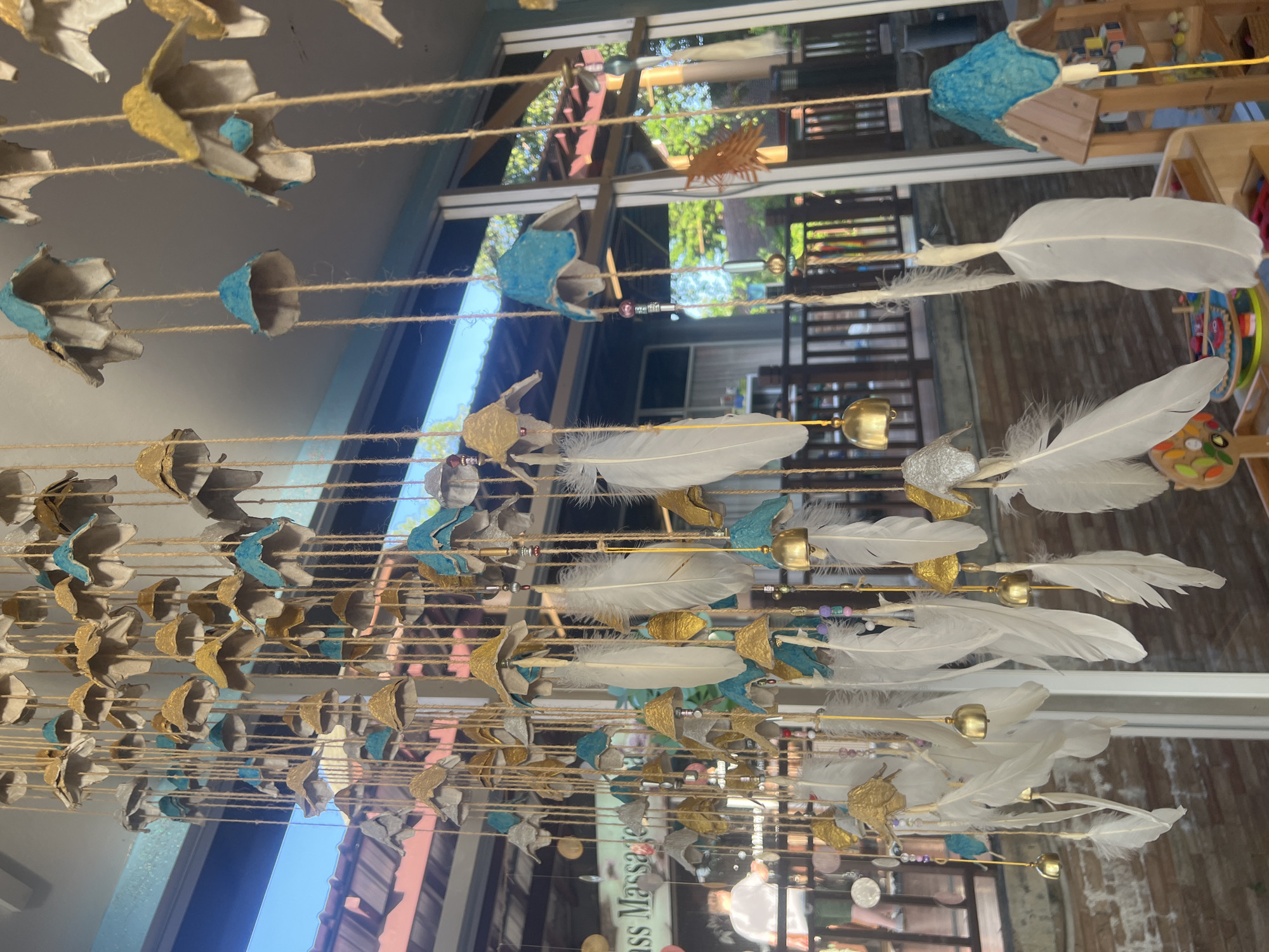 This image shows a close-up of a hanging mobile decoration composed of numerous individual elements suspended from twine. The elements appear to be crafted from repurposed egg cartons, some of which are painted in blue and gold, and others left in their natural beige color. These egg carton pieces are shaped like inverted flowers or bells. Interspersed among these are white feathers of various sizes, some appearing fluffy and others more delicate. There are also small gold-colored bells attached to some strings. In the background, visible through a large window or open doorway, is an outdoor setting with wooden railings and greenery, suggesting a tropical environment. A portion of a sign that reads "ass Massage" is visible on the left side. A person wearing a white t-shirt and green shorts is partially visible in the background, standing near a railing. The overall scene suggests an outdoor space, possibly a shop or a wellness center, in a tropical location like Ko Samui District, Thailand.