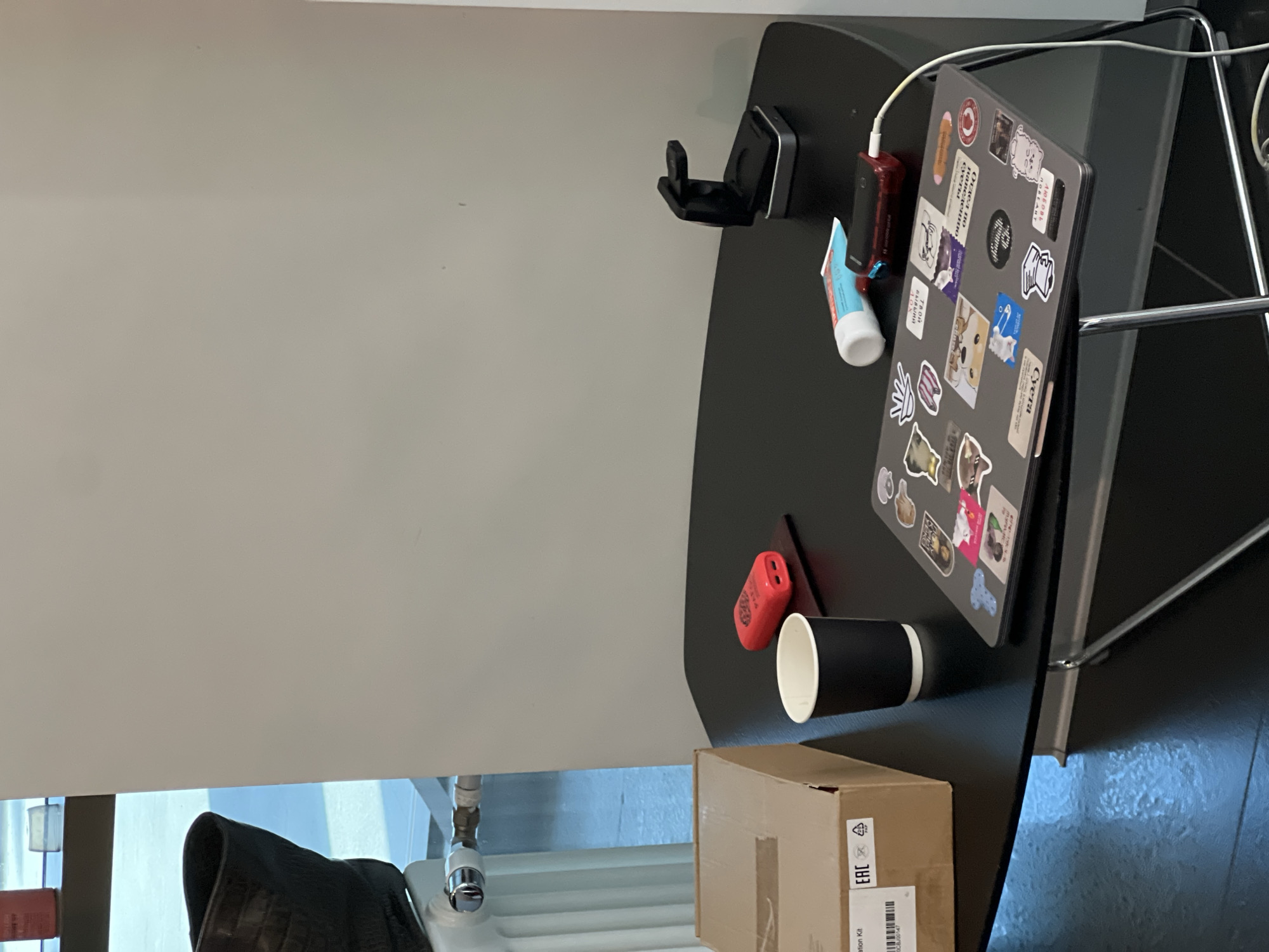 The image depicts an indoor scene, likely in a modern commercial or public space, situated in Moscow, Russia. The specific location is not precisely identifiable beyond the city.
In the foreground, a black, irregularly hexagonal table with chrome legs occupies the lower half of the frame. On the table, from left to right:
* A brown cardboard box is positioned on the far left. It features an "EAC" (Eurasian Conformity) mark, a "PAP" recycling symbol, and a barcode. Partially visible text suggests "lation Kit".
* A black disposable coffee cup with a white interior rim sits to the right of the box.
* A red portable charger or power bank rests on a dark coaster or mat.
* A closed, silver-grey laptop, heavily covered with various stickers, some featuring Cyrillic text, is placed centrally. A white charging cable extends from the laptop's right side, leading out of frame downwards.
* Next to the laptop, a light blue and white tube, possibly containing cream or toothpaste, is visible.
* Further to the right, a black charging stand or phone holder with a metallic base is positioned.
In the mid-ground, to the left of the table:
* A white panel radiator with chrome valves is mounted against a light grey wall.
* A dark, possibly black, bag or object is partially visible on top of the radiator.
* Behind the radiator, a large window allows natural light to illuminate the scene, revealing a bright, possibly snow-covered or icy, outdoor ground.
The background consists primarily of a plain light grey wall, with a small portion of a window visible in the upper left corner. The floor is dark and reflective, mirroring light from the window. No individuals are present in the scene.