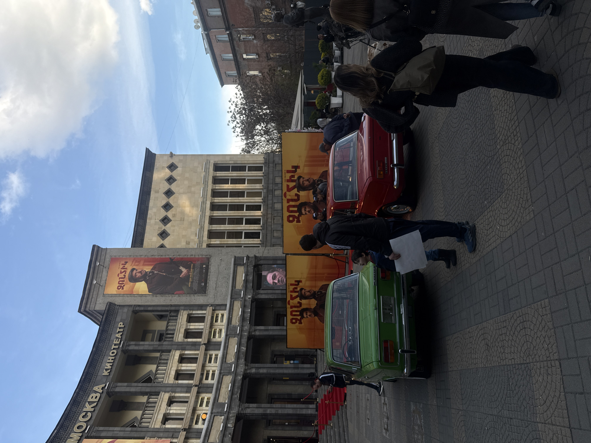 The image captures a scene in Yerevan, Armenia, in front of the Moscow Cinema building. The cinema's facade is visible, adorned with posters in Armenian. Two classic Lada cars, one red and one green, are parked in front of the building. Several people are present, including pedestrians walking by and individuals interacting with the vehicles. A red carpet leads to the entrance of the cinema. The sky is blue with some clouds.